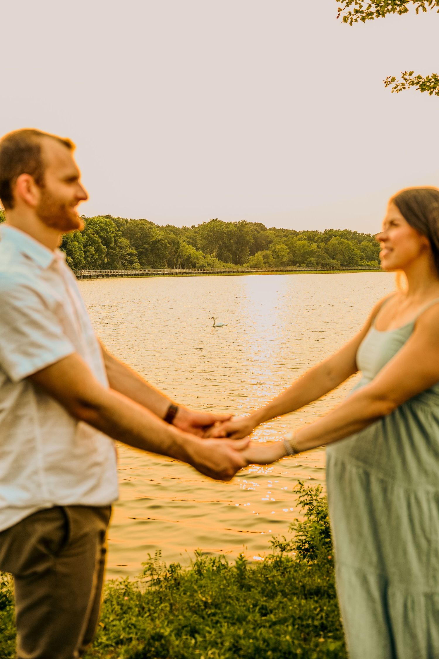 Couple holding hands by a golden lake at sunset with a swan swimming in the background and lush green trees.