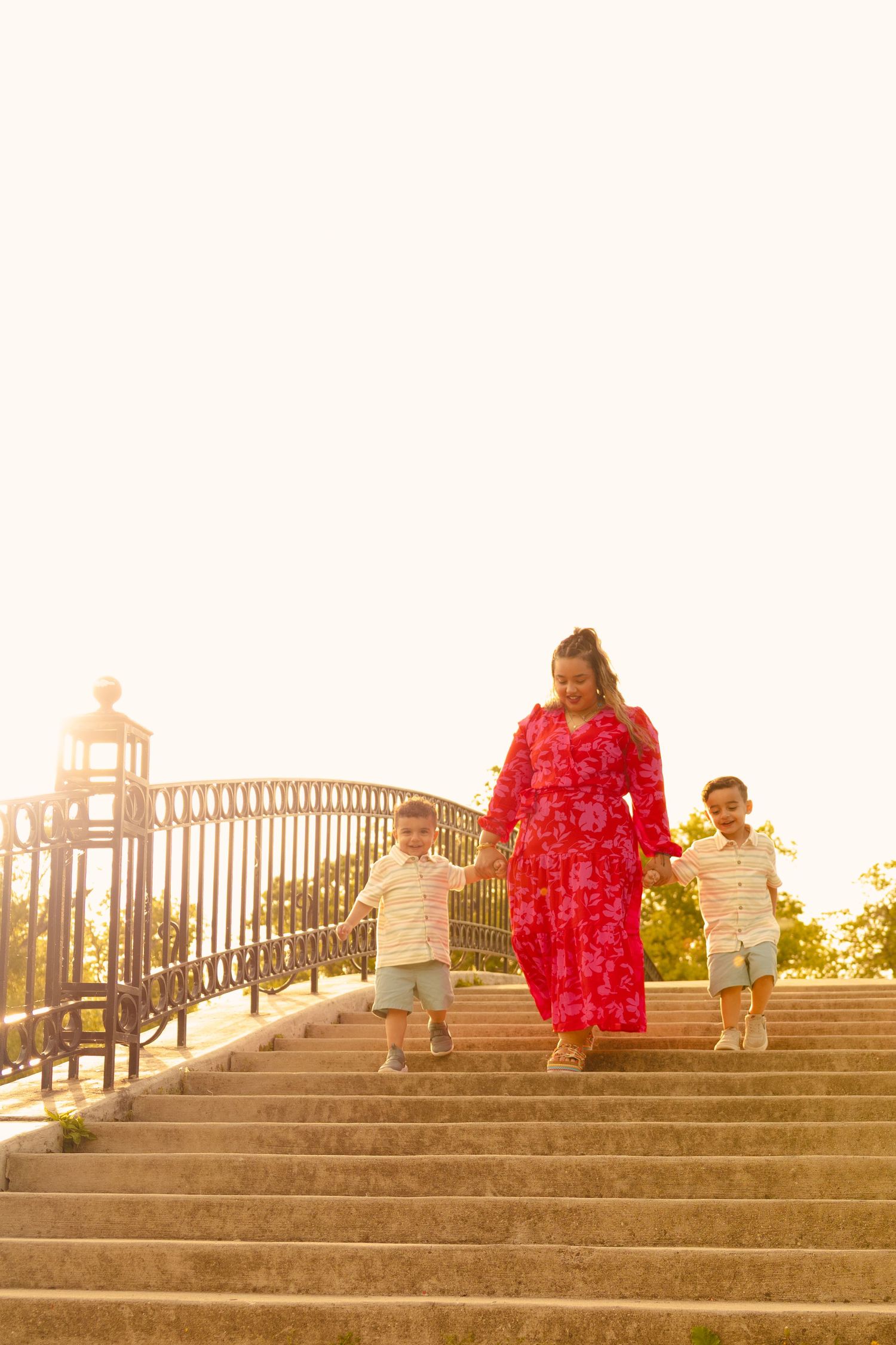 A mother in a red floral dress walks down sunny outdoor steps holding hands with her two young sons.