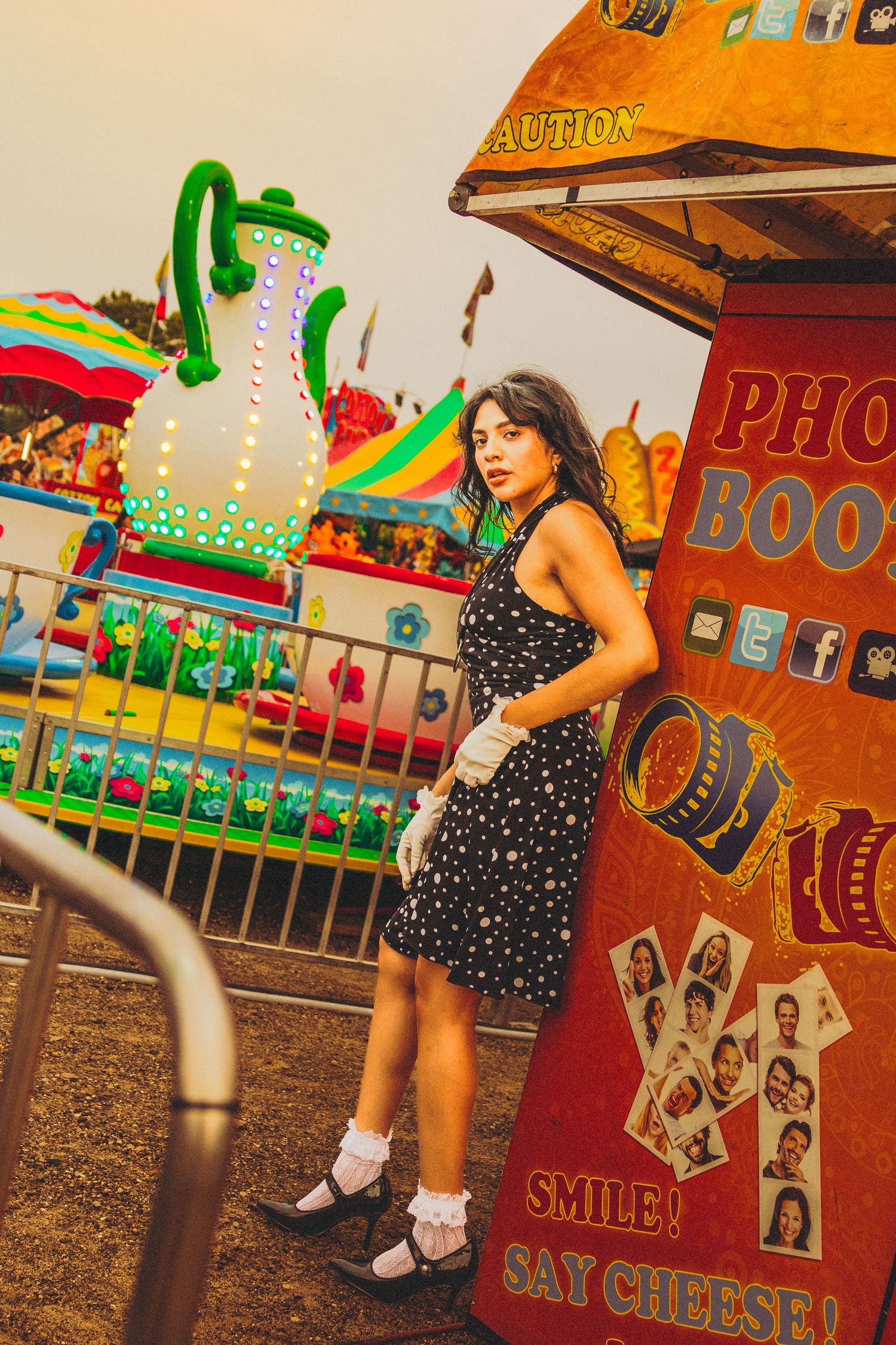 Young woman in polka dot dress posing by a photo booth at a colorful carnival fairground.