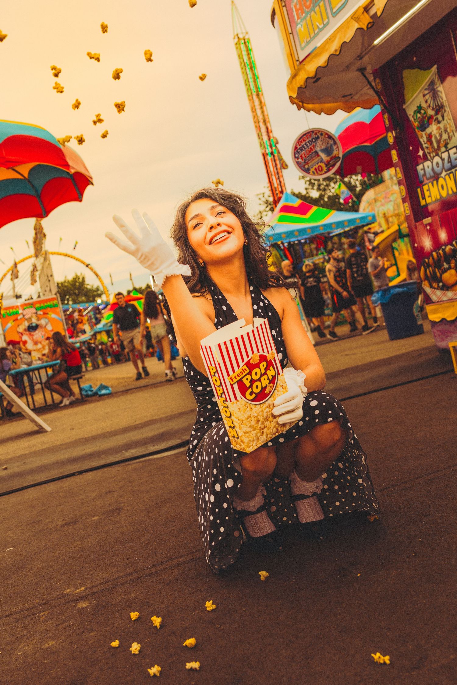 Young woman in polka dot dress tossing popcorn at a colorful carnival fair, smiling joyfully while holding a popcorn bucket.
