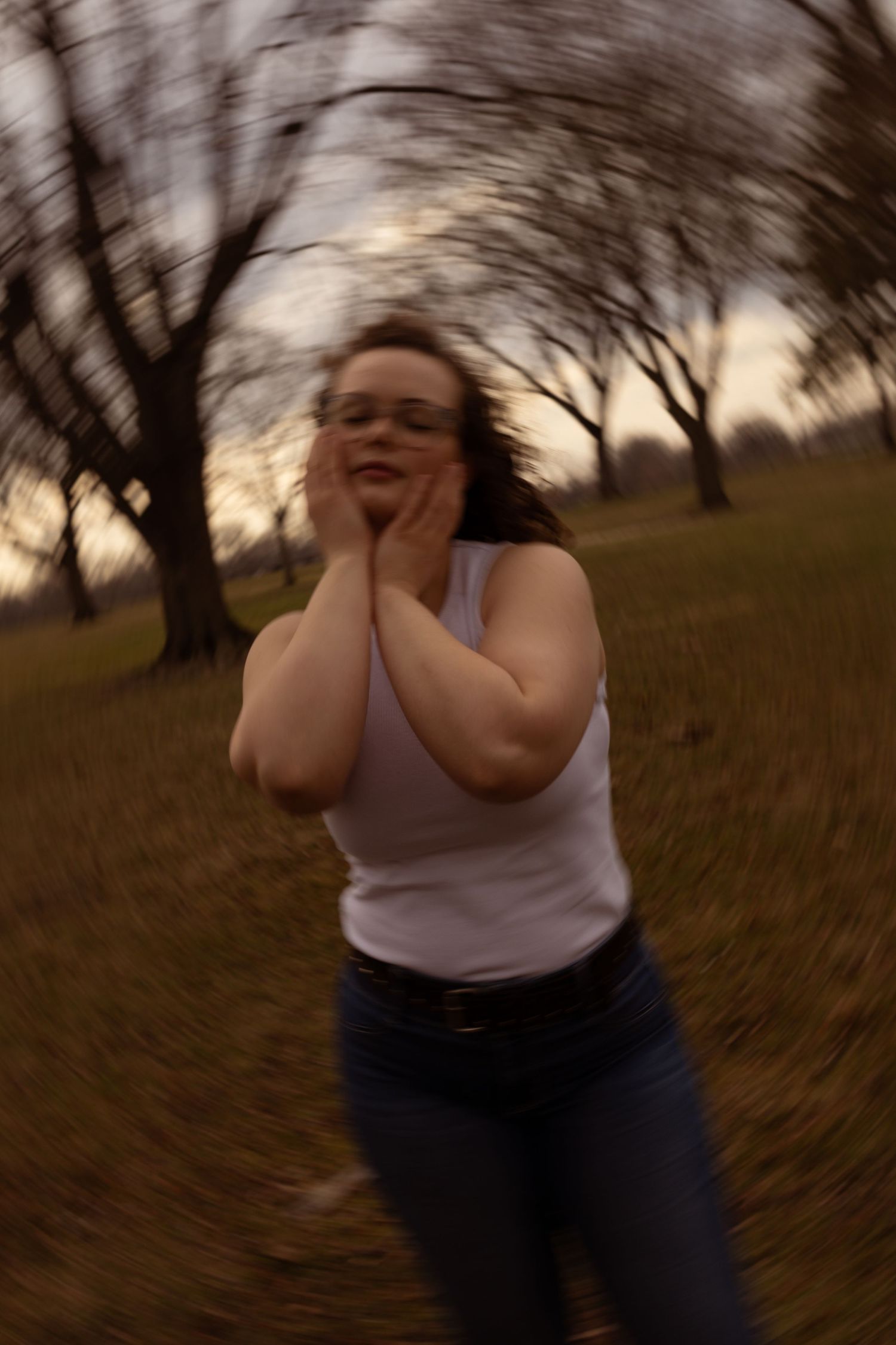 A young woman in a white tank top and jeans poses outdoors in a park with bare trees at dusk.