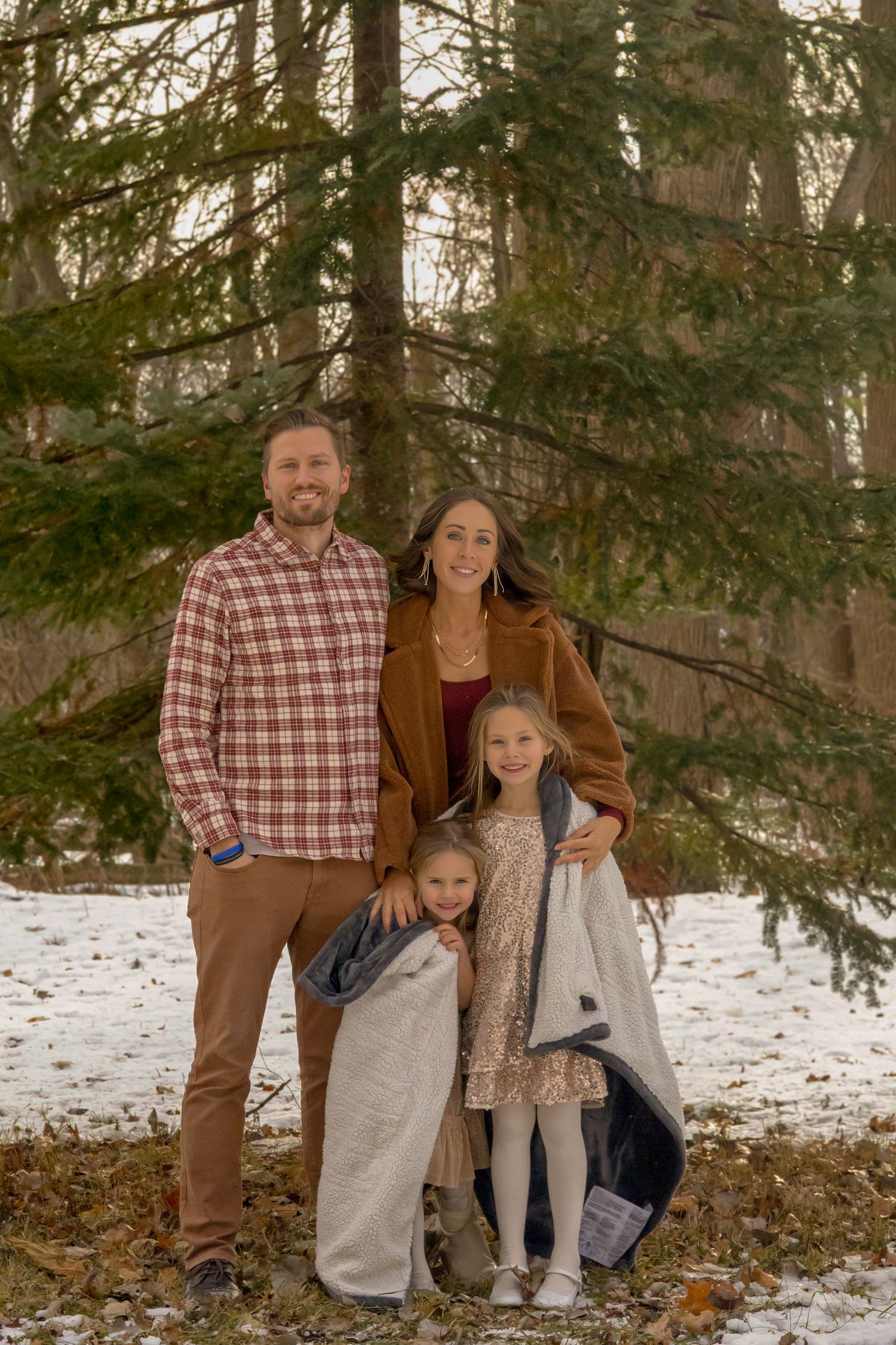 Family of four posing outdoors in winter, surrounded by snow-dusted evergreen trees, wearing warm fall and winter clothing.