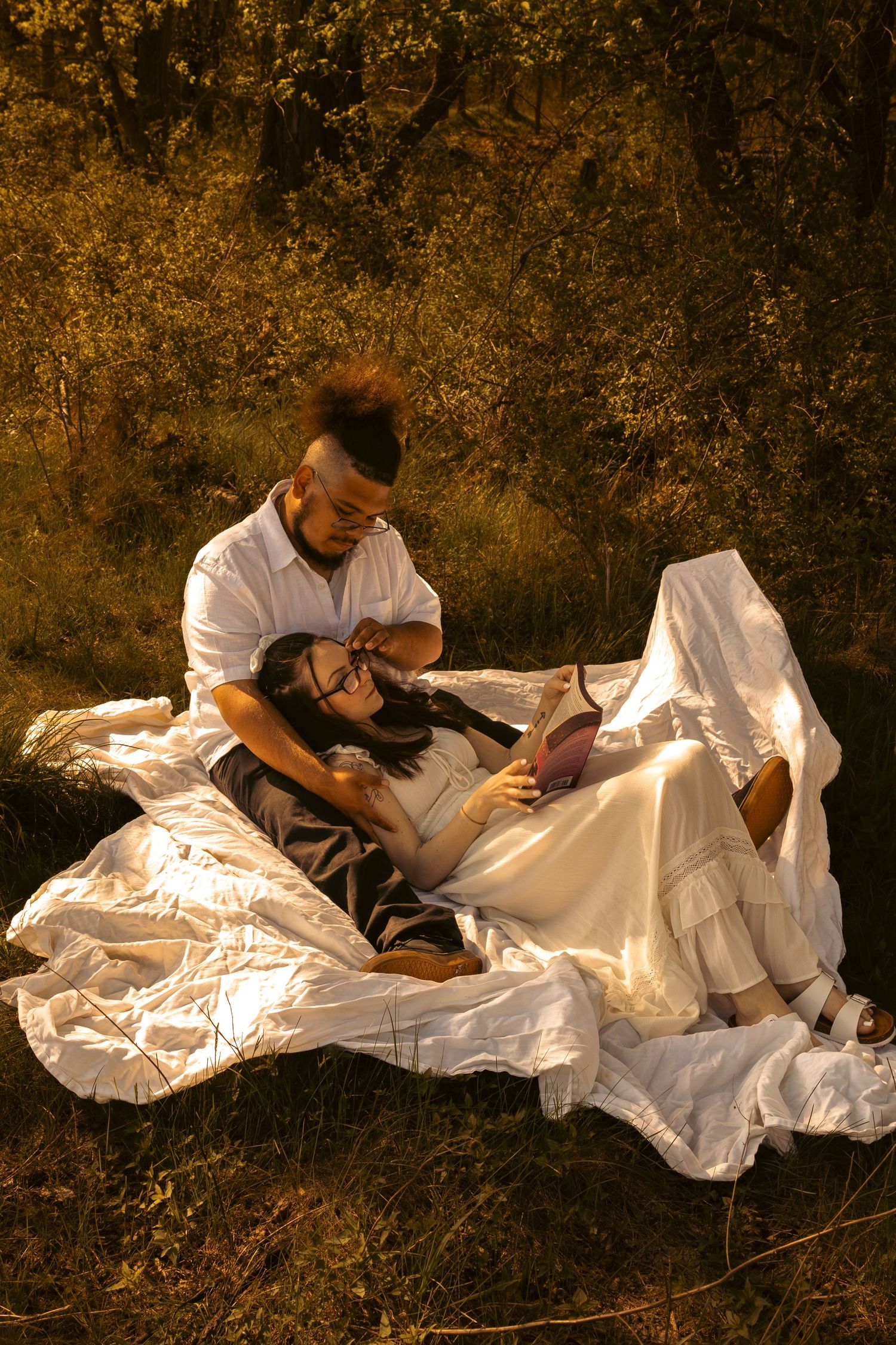 Couple relaxing on white blanket in sunlit forest, woman lying in mans lap reading a book outdoors.