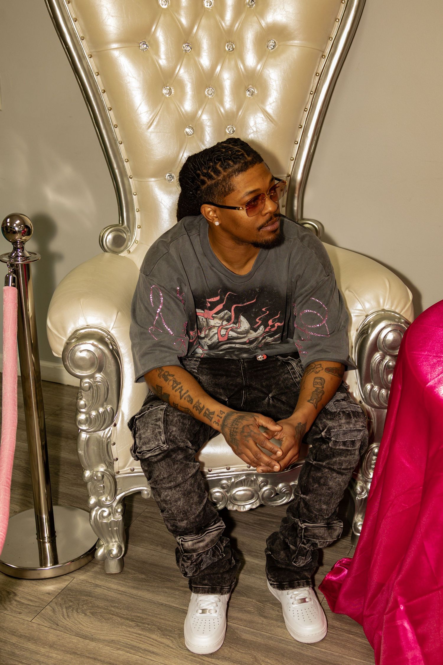 Young man with braids sits in a silver tufted throne chair wearing graphic tee, dark jeans, and white sneakers.