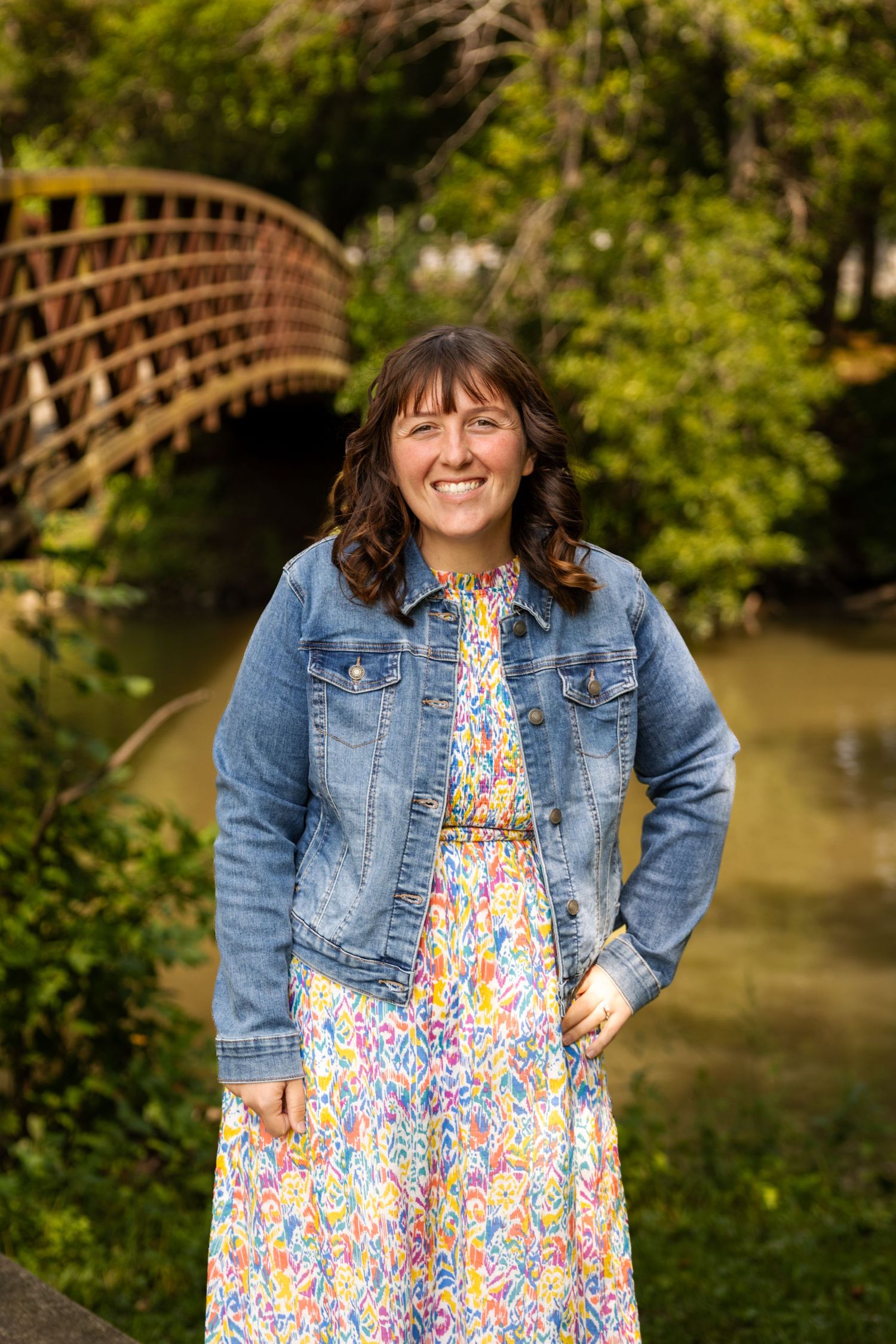 Smiling woman in floral dress and denim jacket standing outdoors near a wooden bridge.