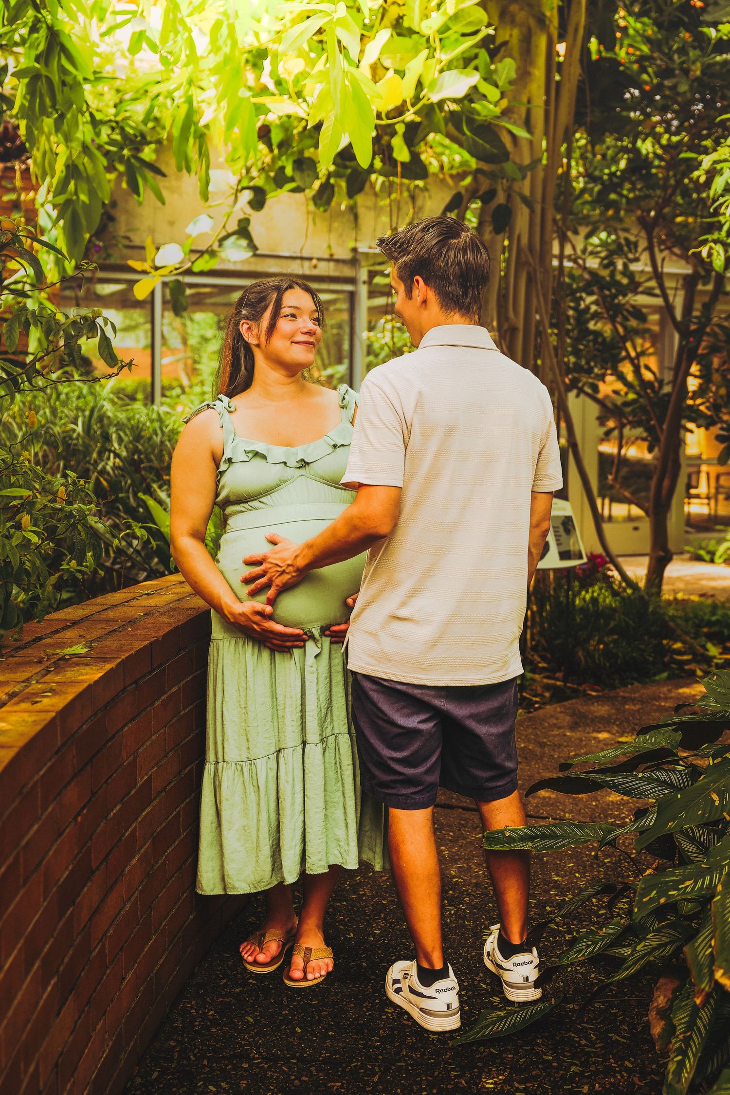 Pregnant woman in green dress holds her belly while talking with a man in a lush garden setting.