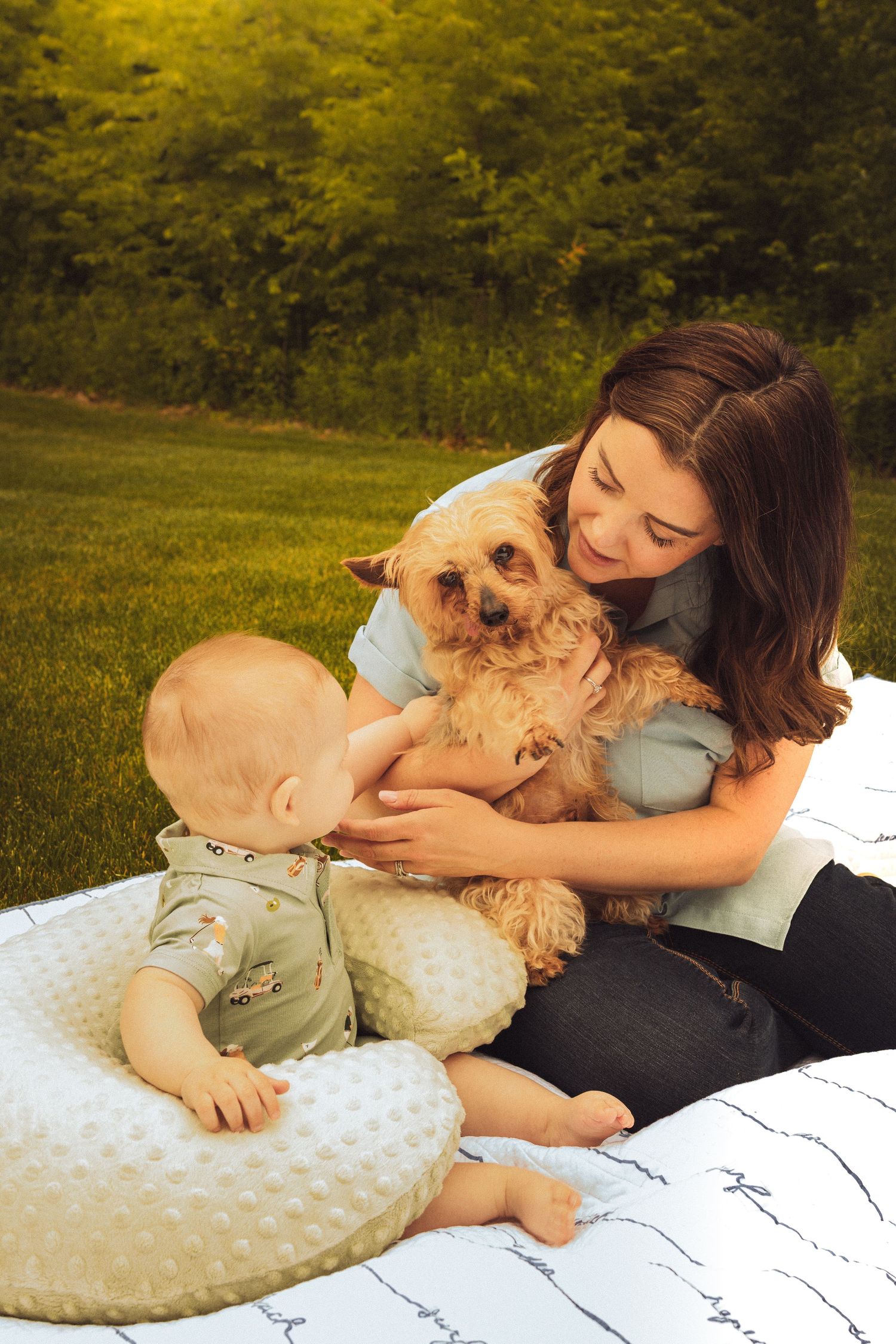 Woman holding small dog while baby reaches out to pet it on a blanket outdoors in a sunny green yard.