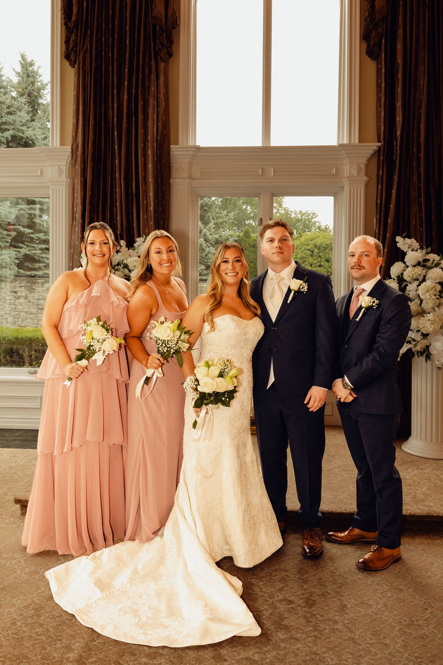 Bride in a lace gown poses with two bridesmaids in blush dresses and two groomsmen in navy suits in an elegant room.