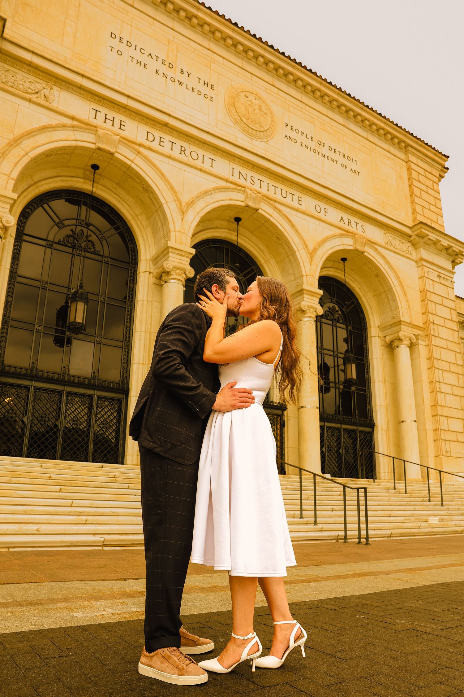 Couple sharing a romantic kiss in wedding attire outside the Detroit Institute of Arts museum.