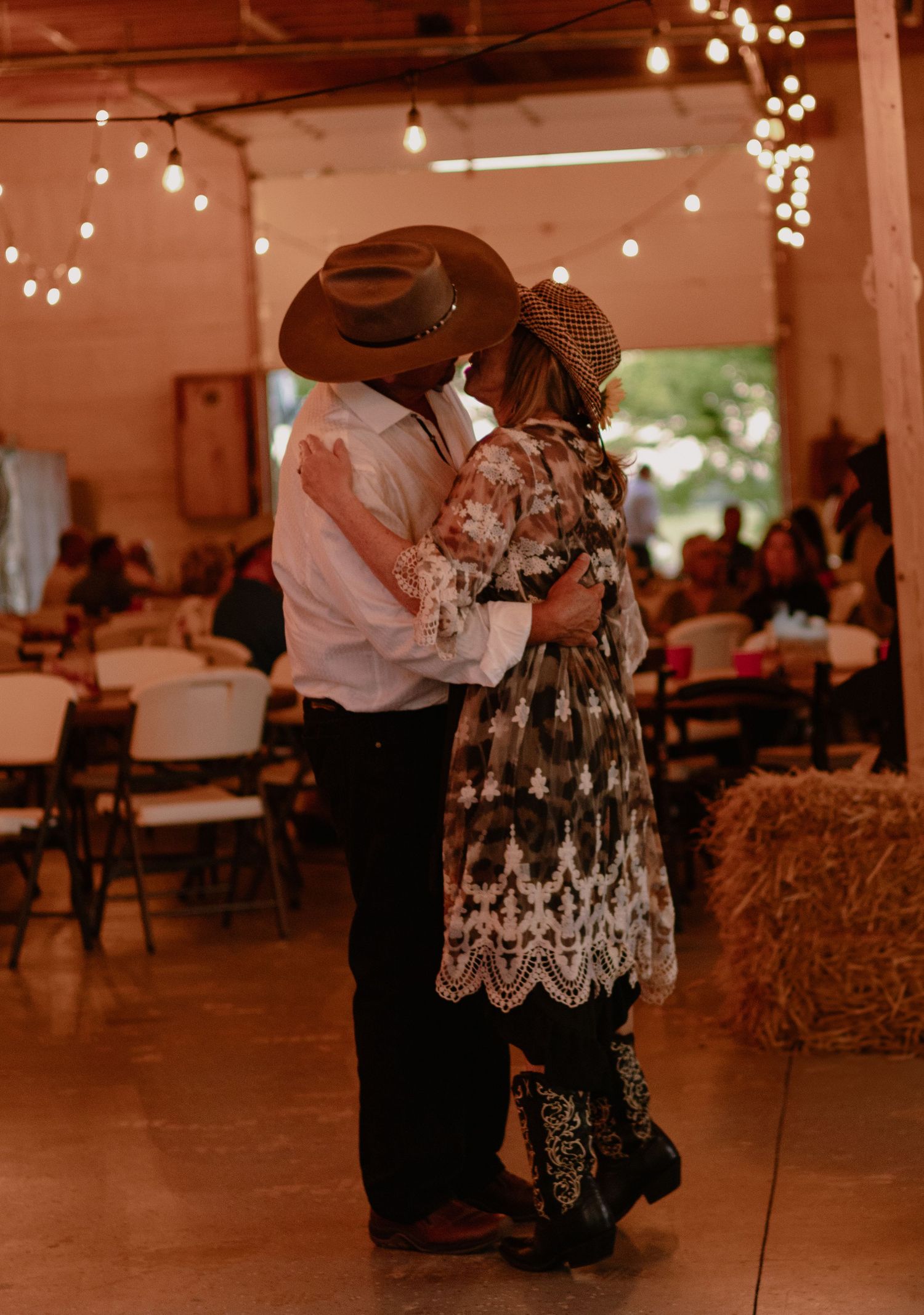 Couple sharing a first dance kiss in cowboy hats at a rustic barn wedding with string lights and hay bales.
