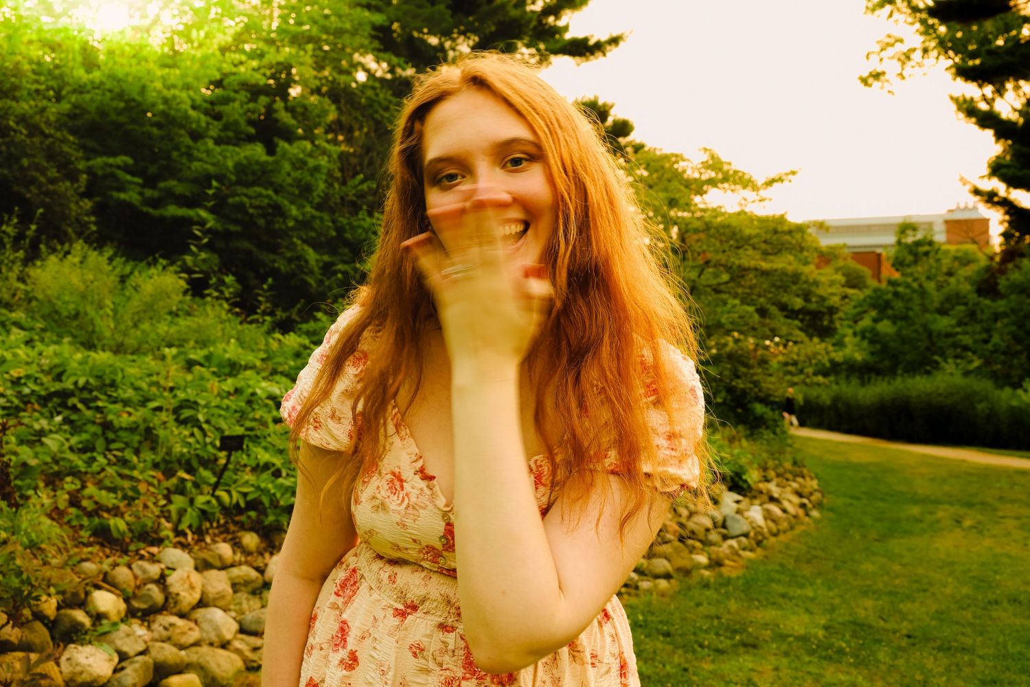 Young woman with long red hair in floral dress laughing outdoors in a lush green garden.