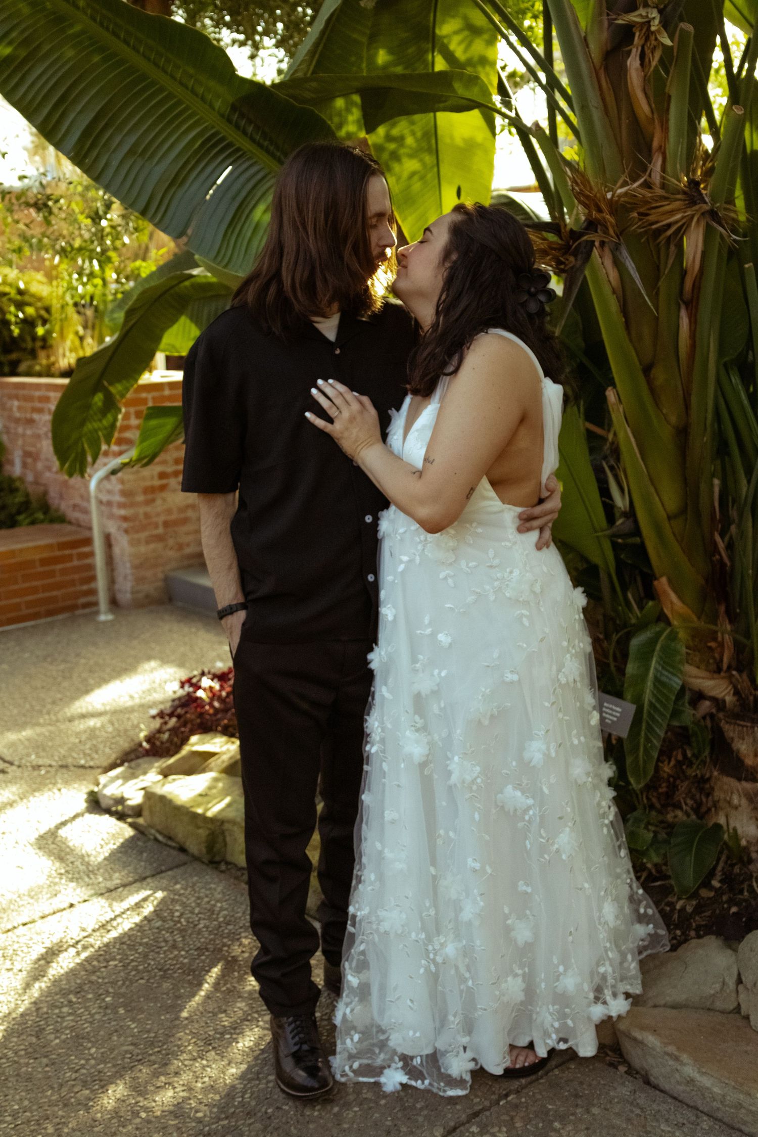 Bride in white floral gown and groom in black suit share a romantic moment outdoors near tropical plants.