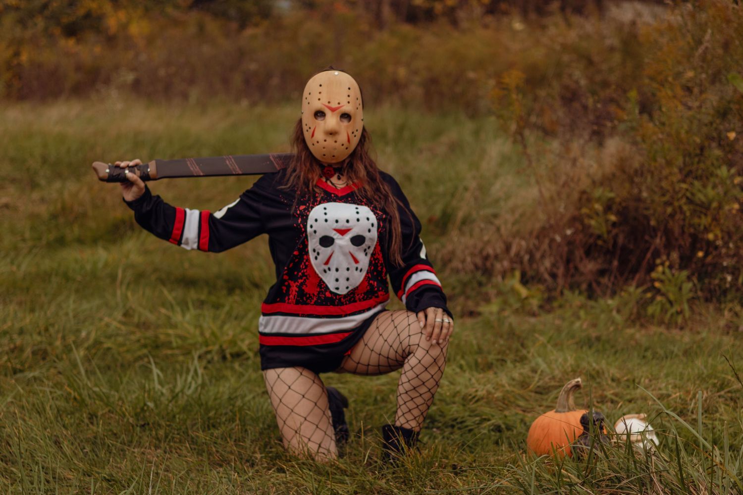 Woman in Jason Voorhees hockey mask and sweater holding machete with pumpkin in autumn field.