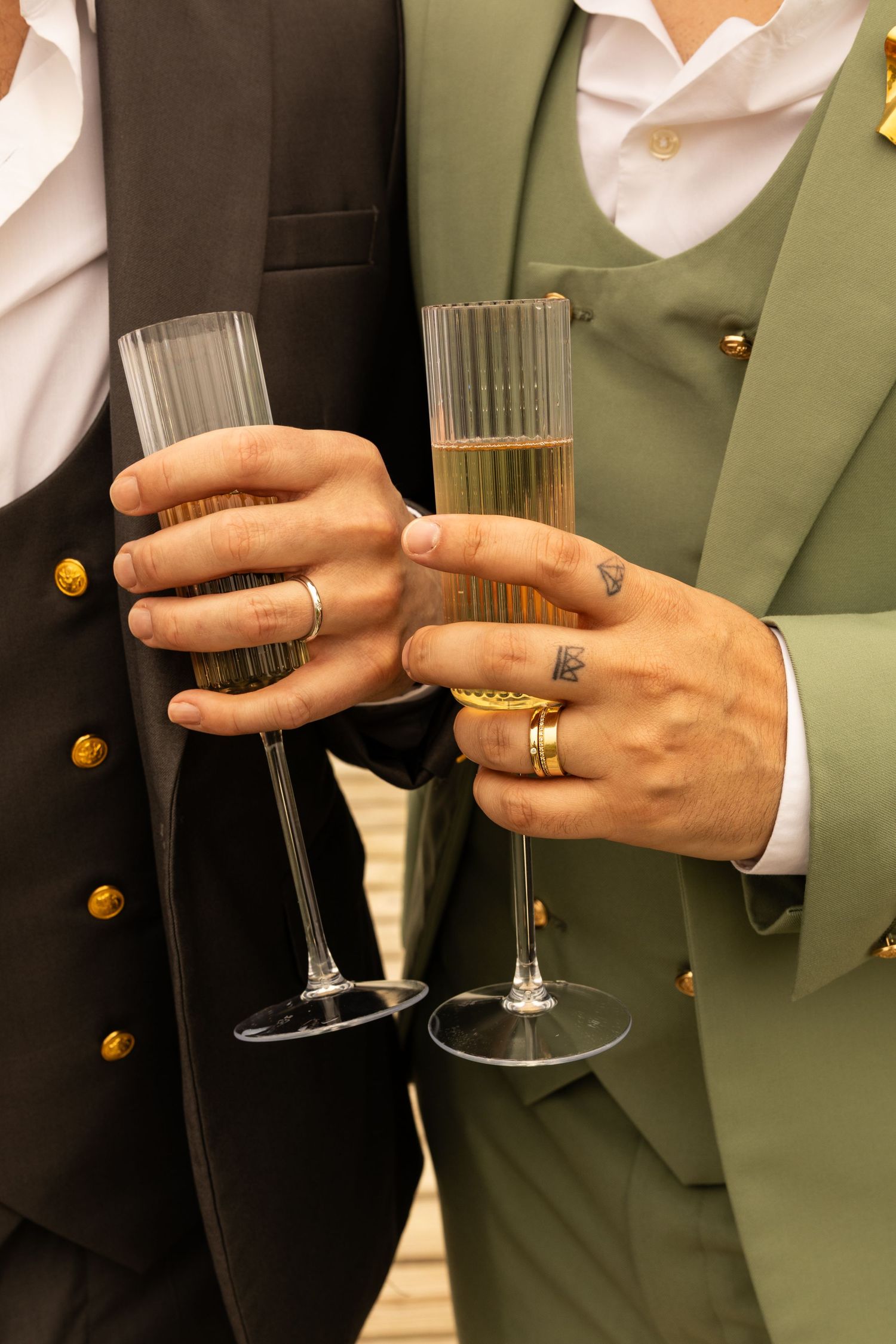 Two grooms in elegant suits toasting with champagne flutes, adorned with rings and tattoos at their wedding celebration.