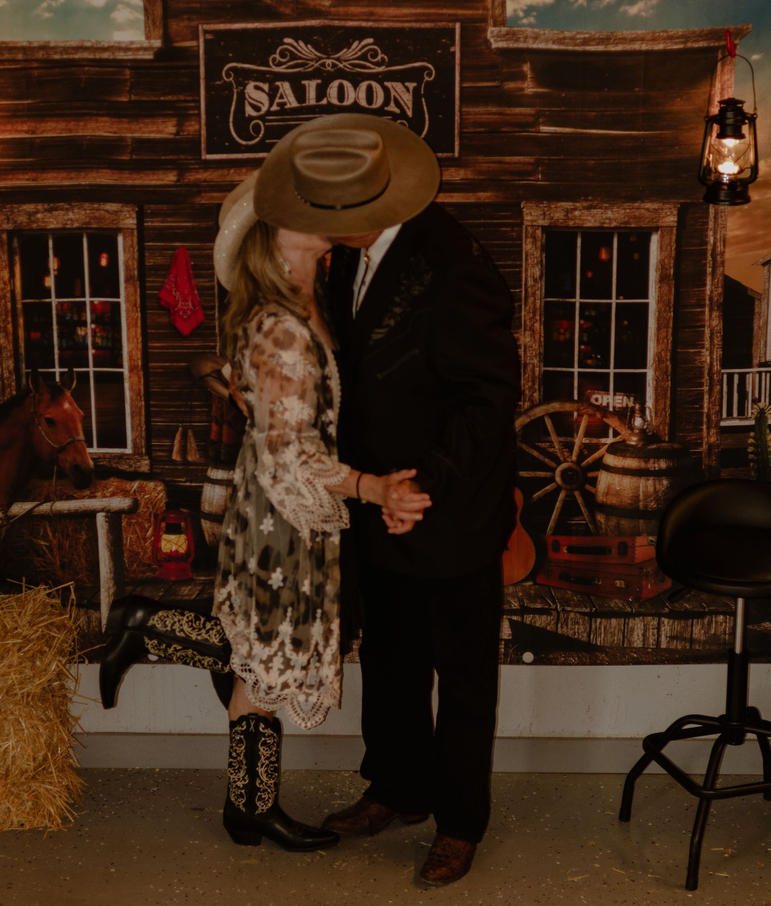 Couple kissing in front of a rustic saloon backdrop with hay bales, a wagon wheel, and Western decor.