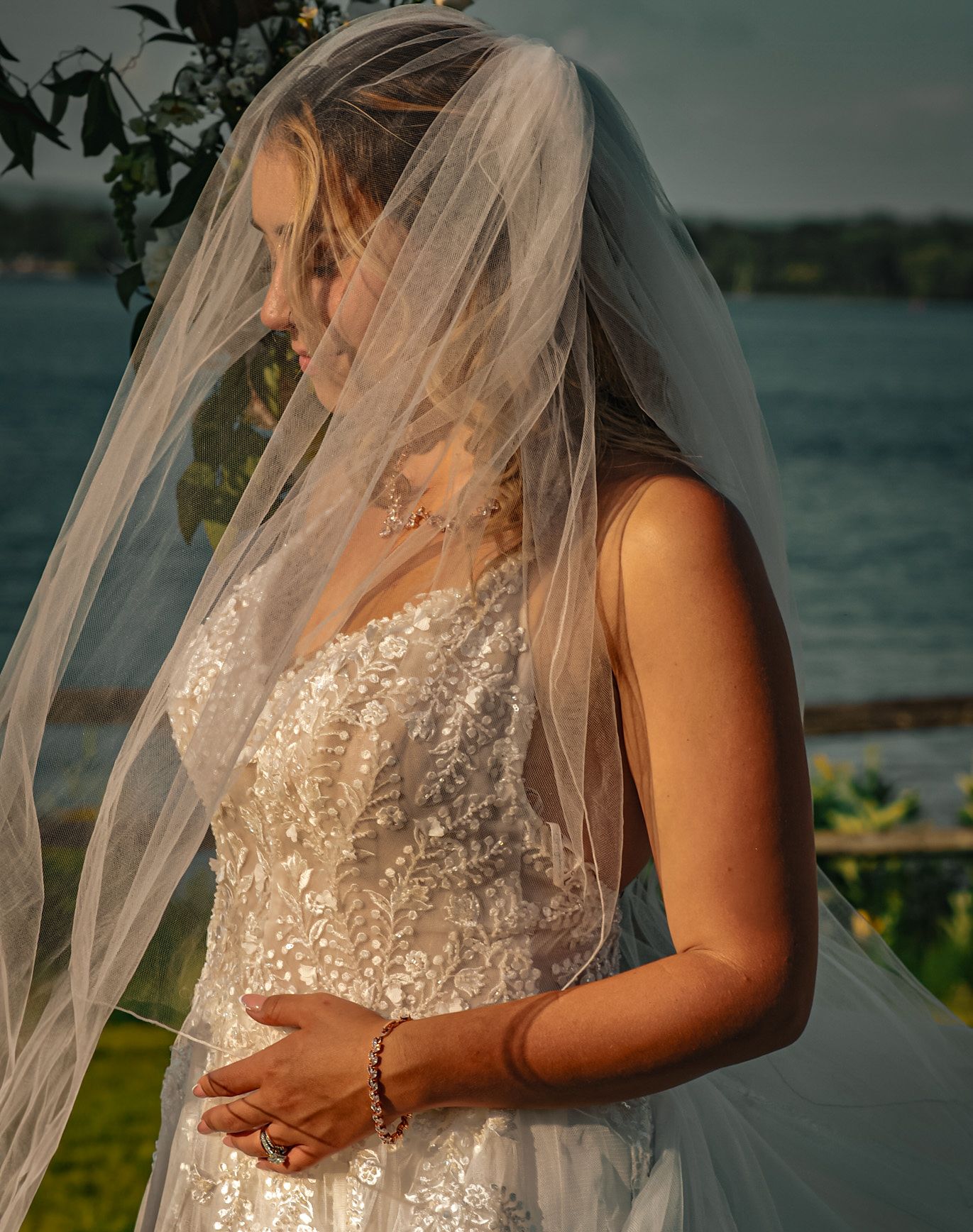 A bride in a detailed lace wedding dress stands by a waterfront at sunset with her veil flowing in the breeze.