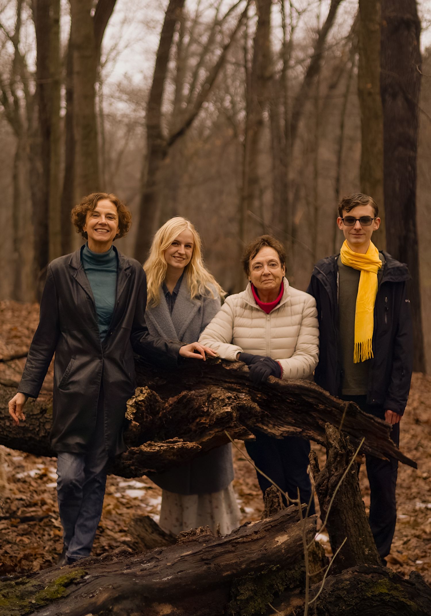 Four people posing together in an autumn forest beside a fallen log, wearing cozy winter coats and scarves.