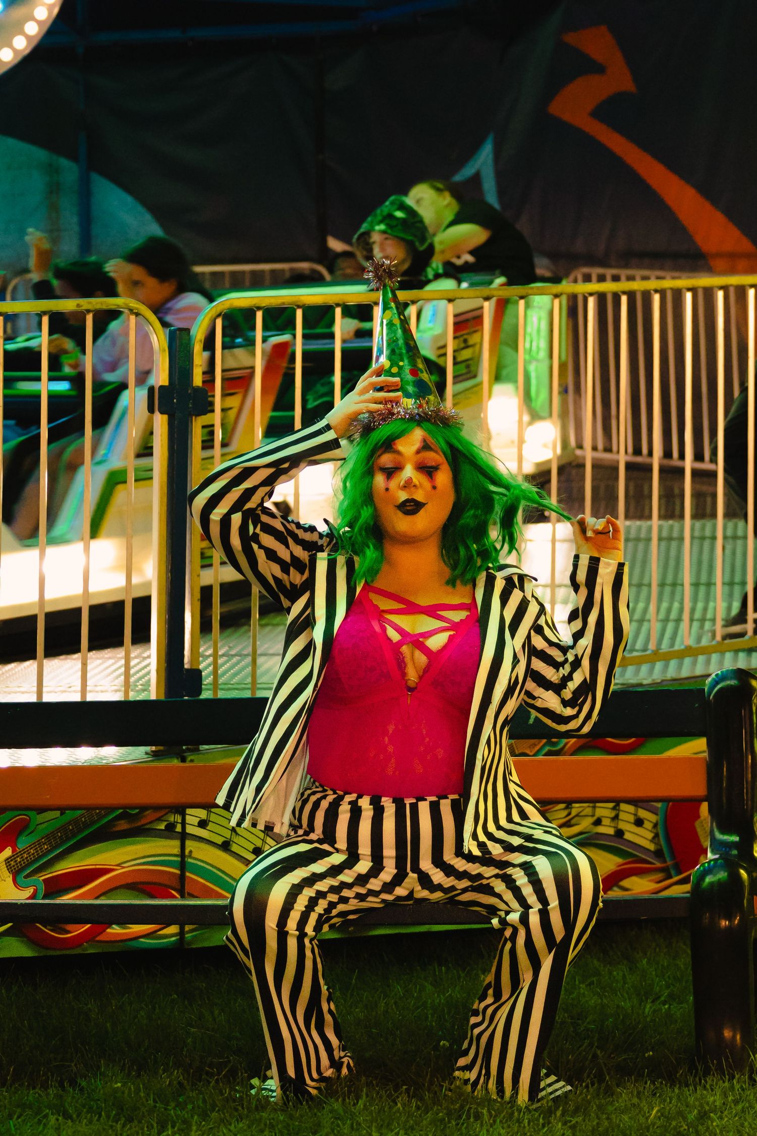 Person in black and white striped suit with green wig and clown makeup poses at a carnival at night.