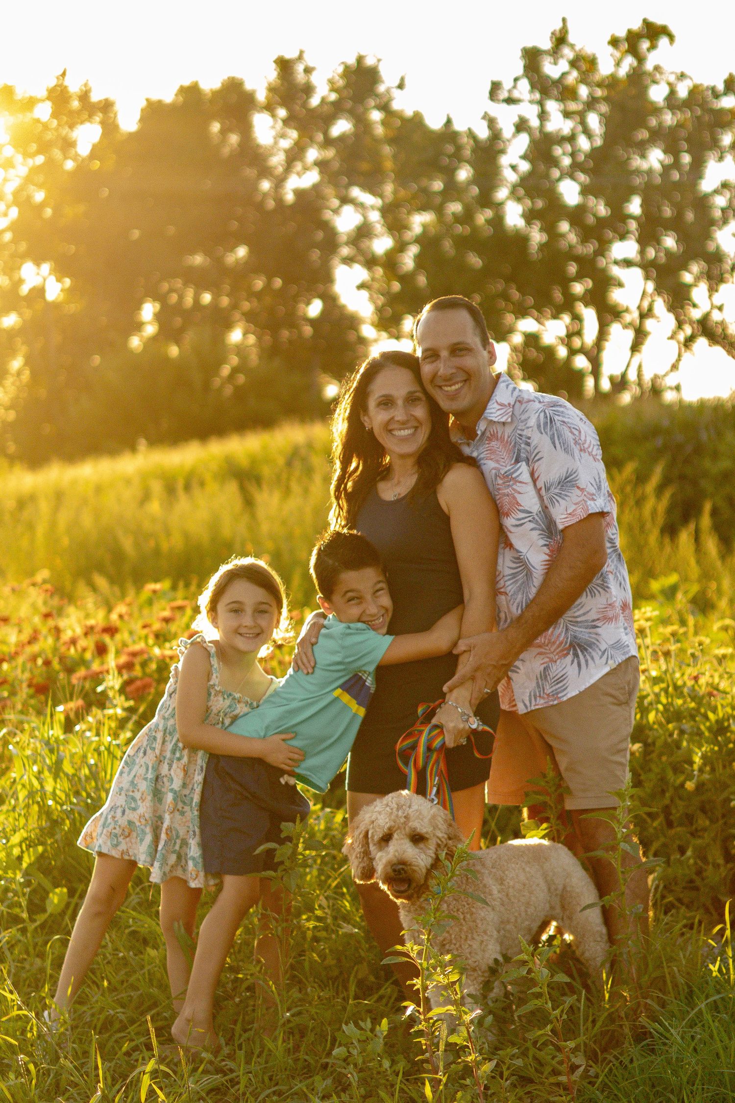 A family poses together with their fluffy dog in a sunlit field during golden hour.