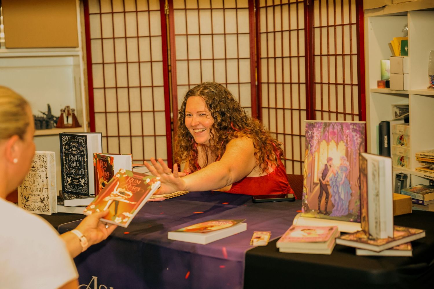 Author smiling at book signing table with novels displayed, engaging with a visitor at an indoor event.