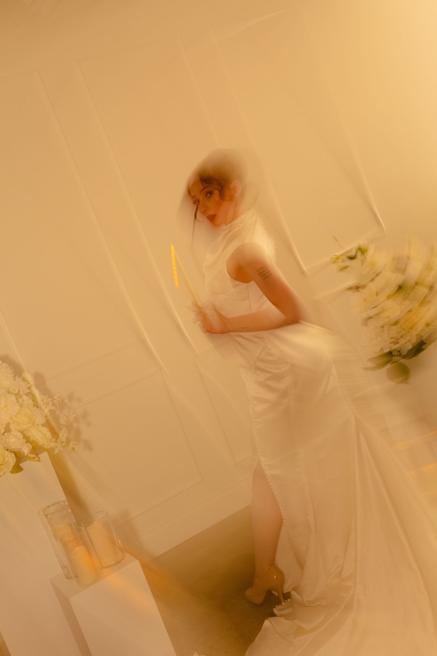 A bride in a white gown and veil holds a candle, surrounded by floral arrangements in a softly lit, dreamy setting.