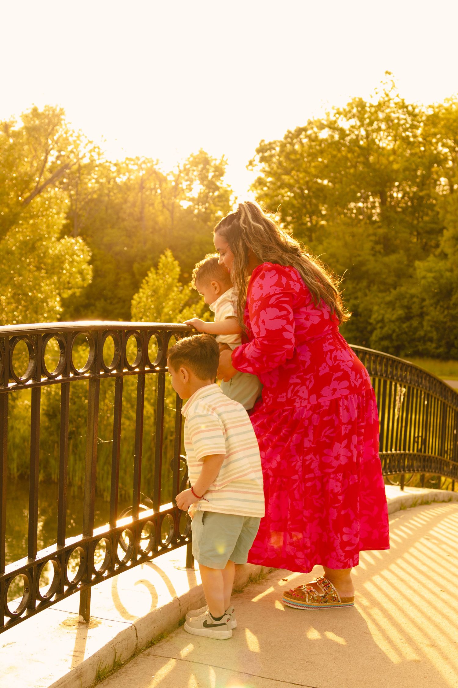 A mother in a red floral dress stands with two young boys on a sunlit bridge overlooking a lush green park.