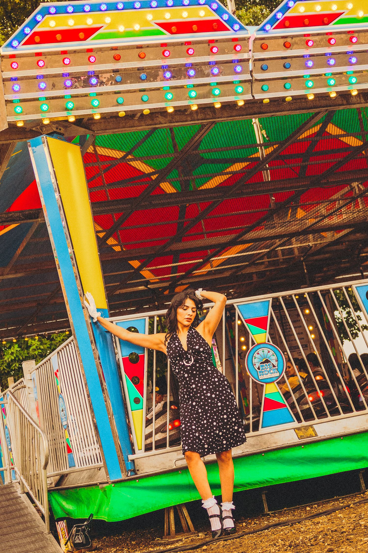 Young woman in polka dot dress posing by a colorful carnival ride with bright lights at an amusement park.