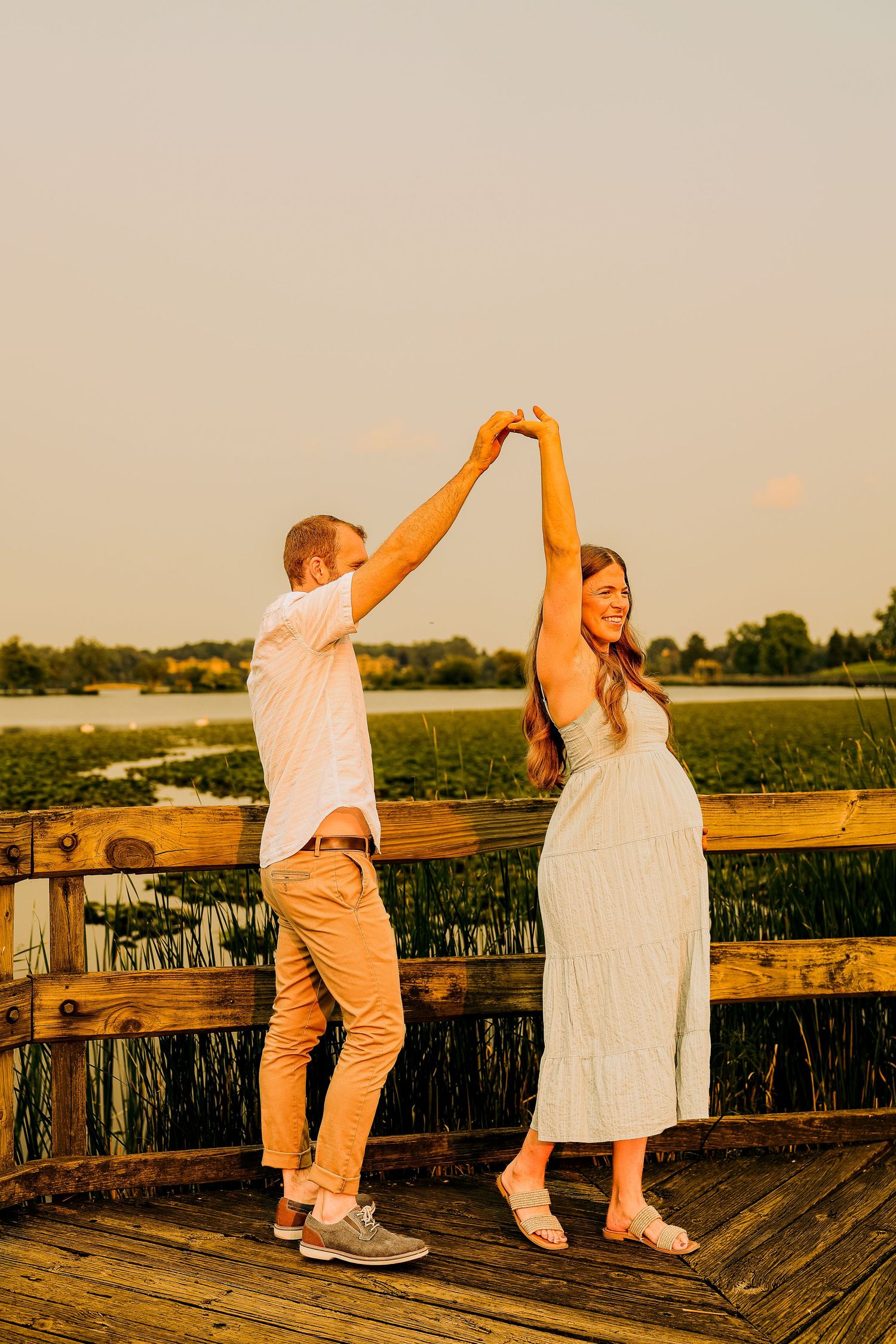Pregnant couple dancing on a wooden dock at golden hour with a lush green marsh and lake in the background.