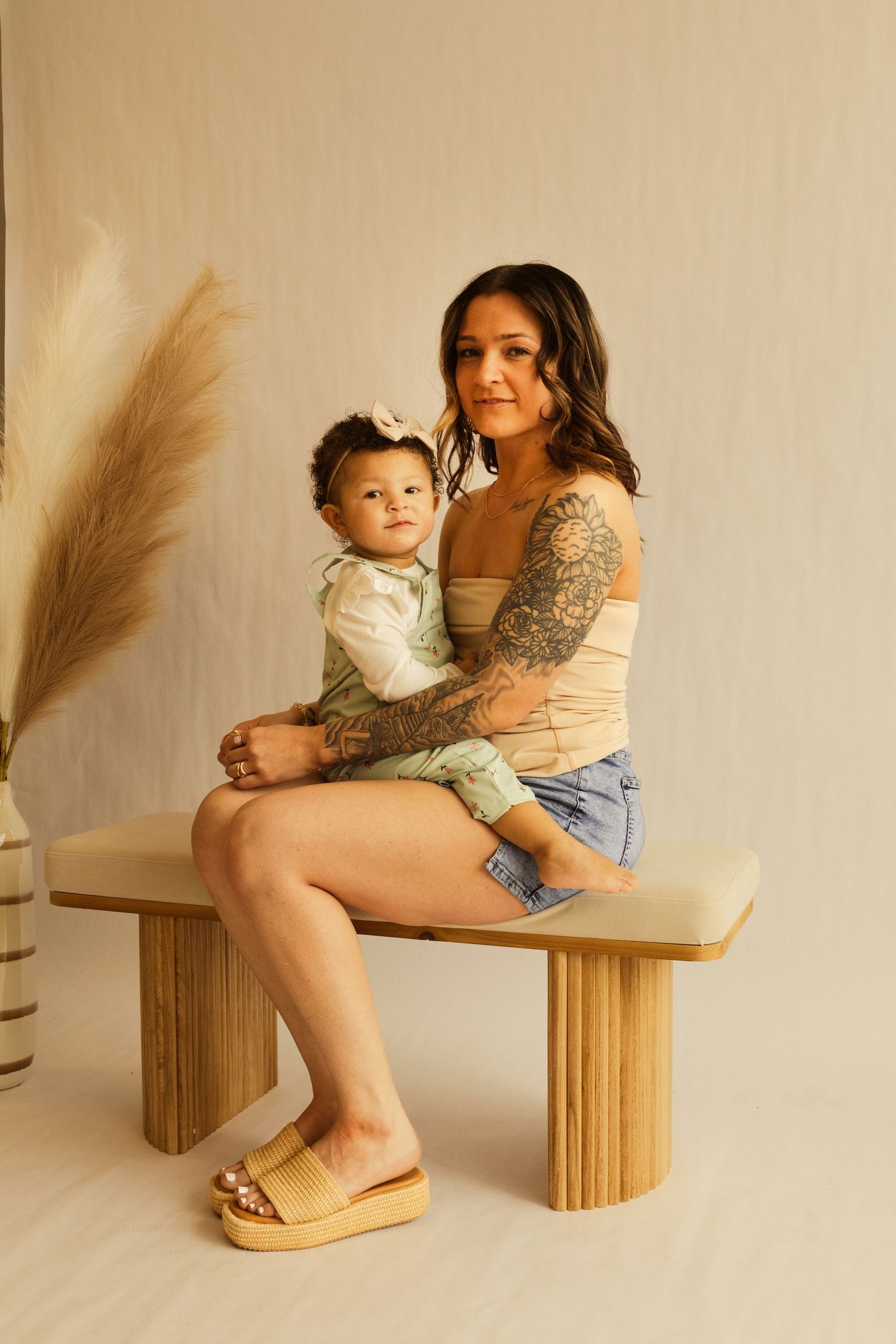 Tattooed mother sitting on a wooden bench holding her baby, wearing denim shorts and a beige top.