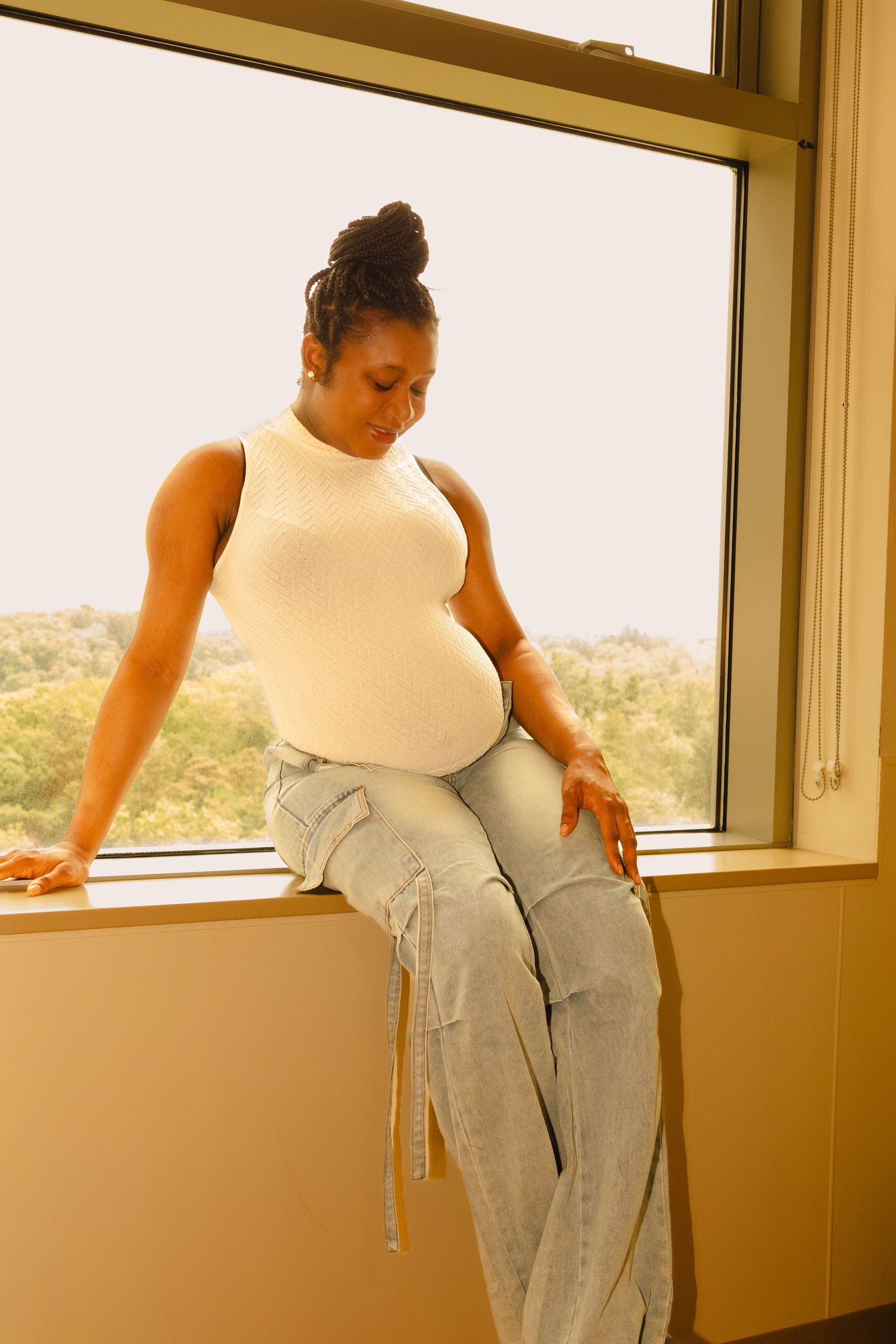 Pregnant woman in white tank top and light jeans sits on windowsill, looking down at her baby bump.