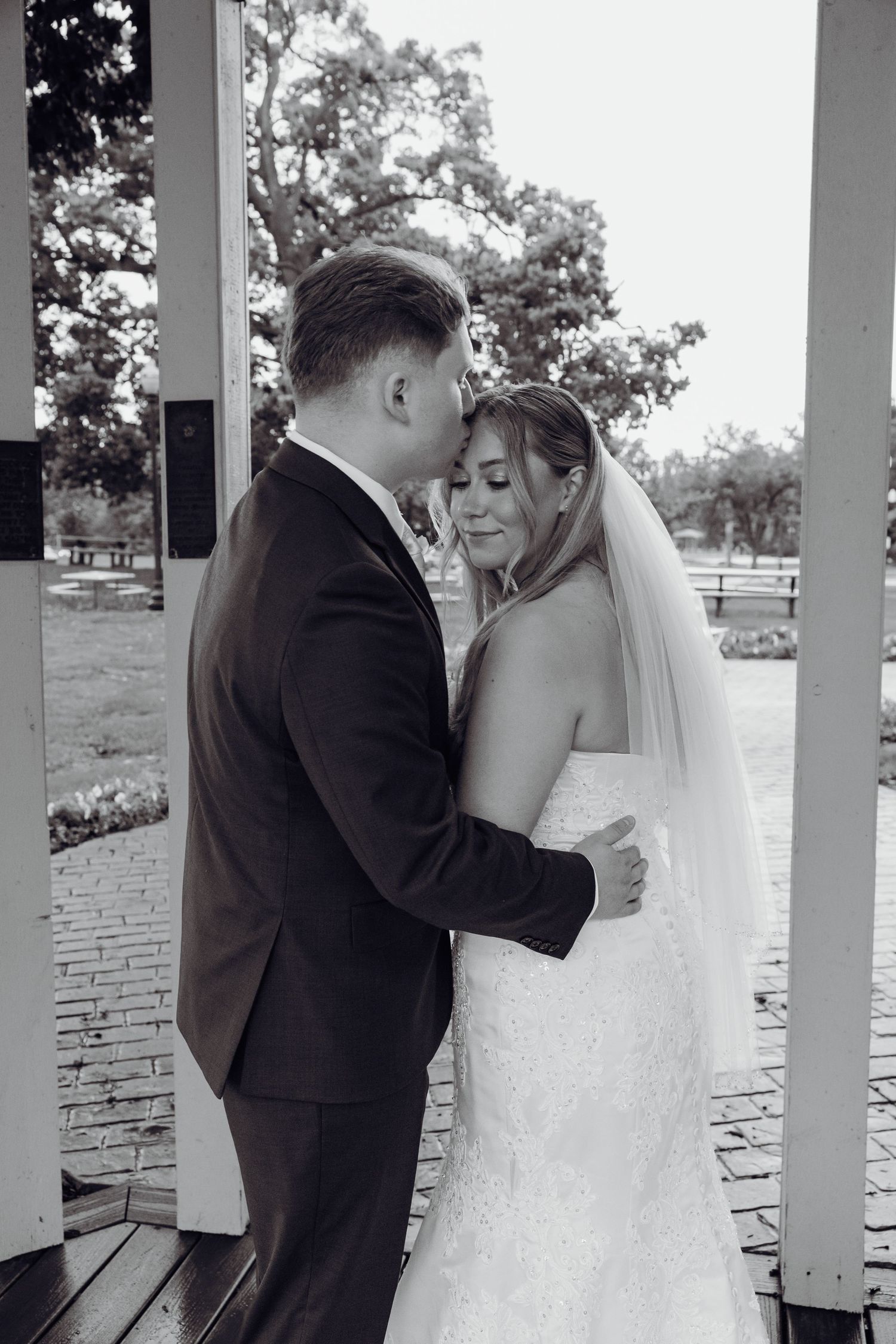 Black and white photo of a groom kissing his bride's forehead as they embrace in a park gazebo, her lace wedding gown and veil flowing.