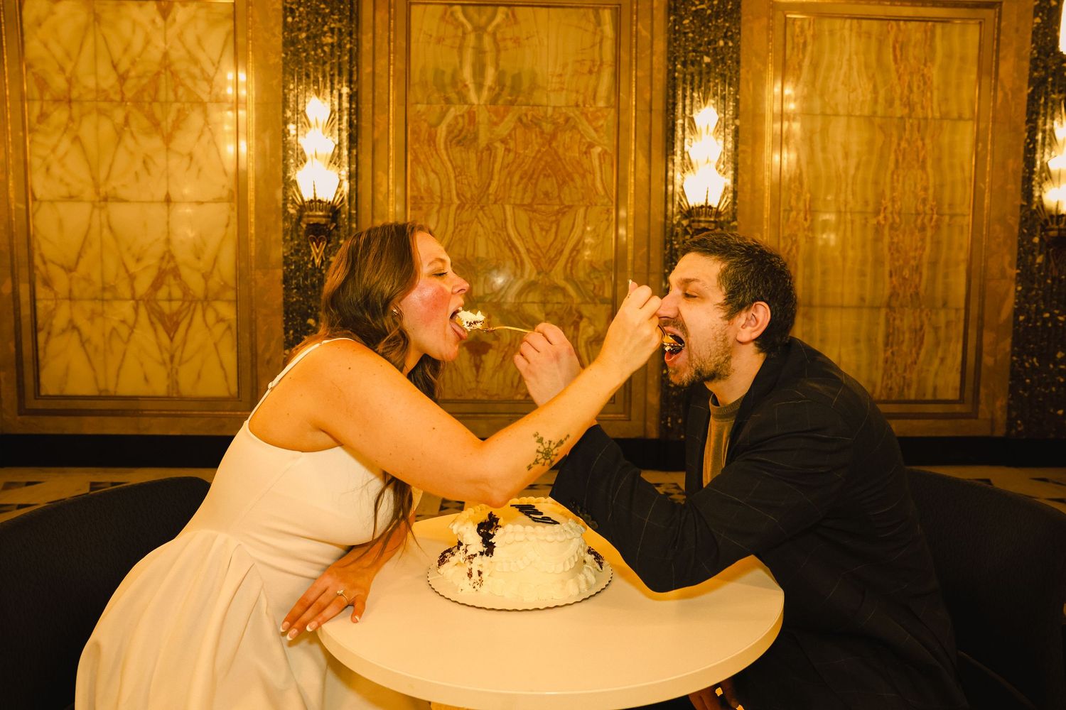 Bride and groom playfully feeding each other wedding cake at a table in an elegant gold-lit reception venue.