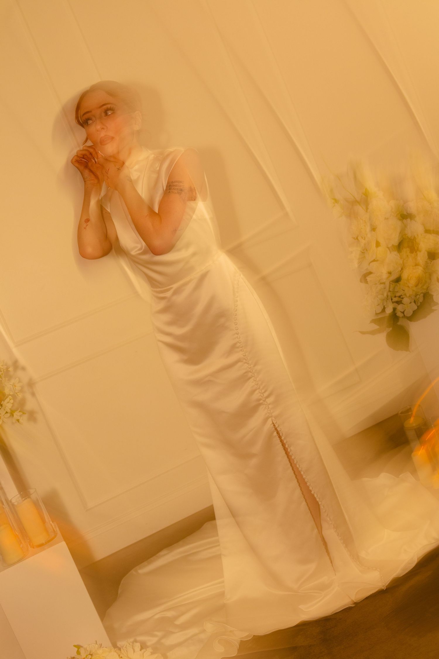 A bride in a white satin gown and veil poses gracefully in a warmly lit room adorned with yellow floral arrangements.