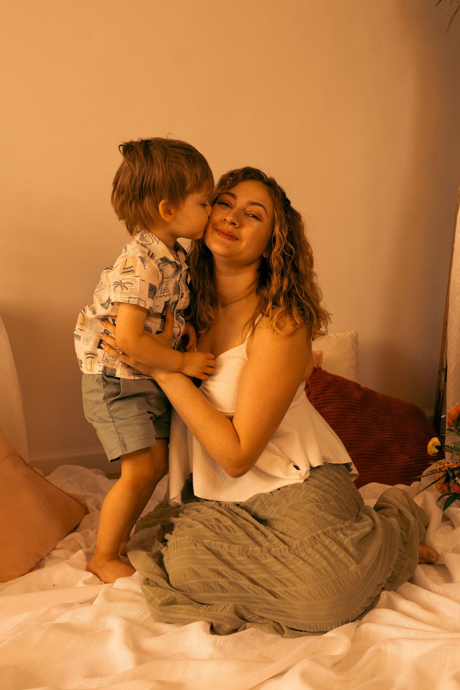 A young boy kisses his smiling mother on the cheek while sitting on a bed in warm lighting.