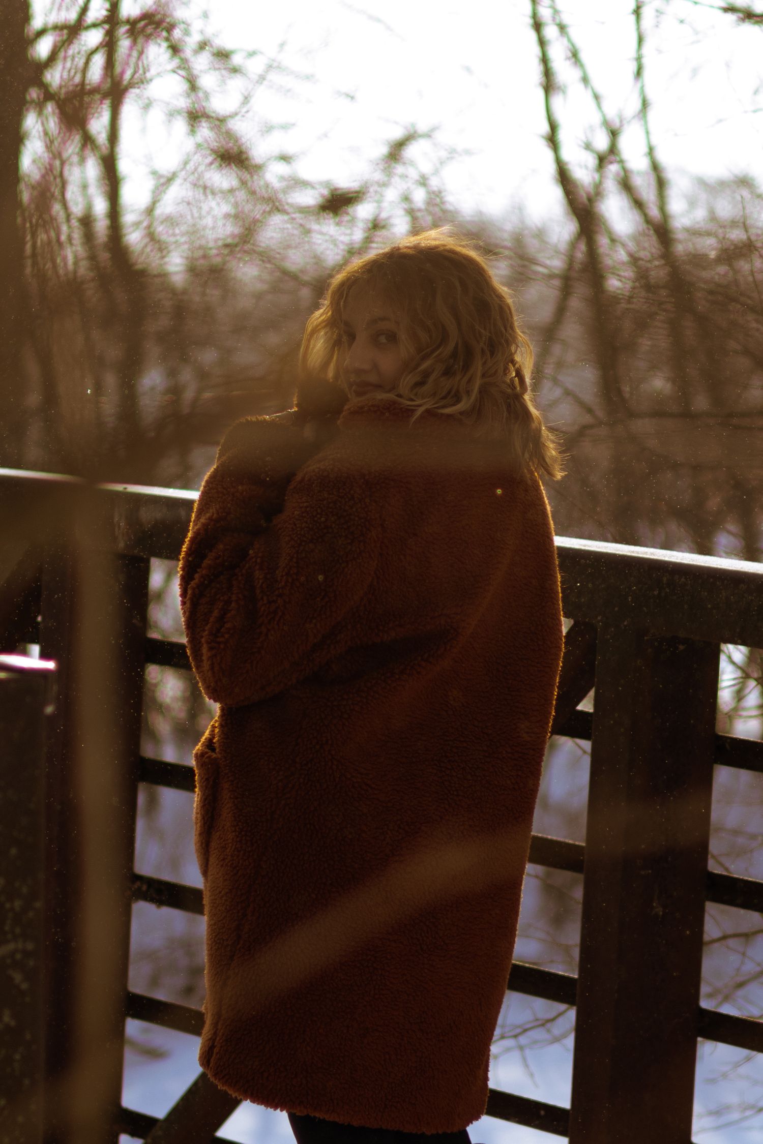 Woman in brown teddy coat standing on a bridge in winter, looking back over her shoulder in golden light.