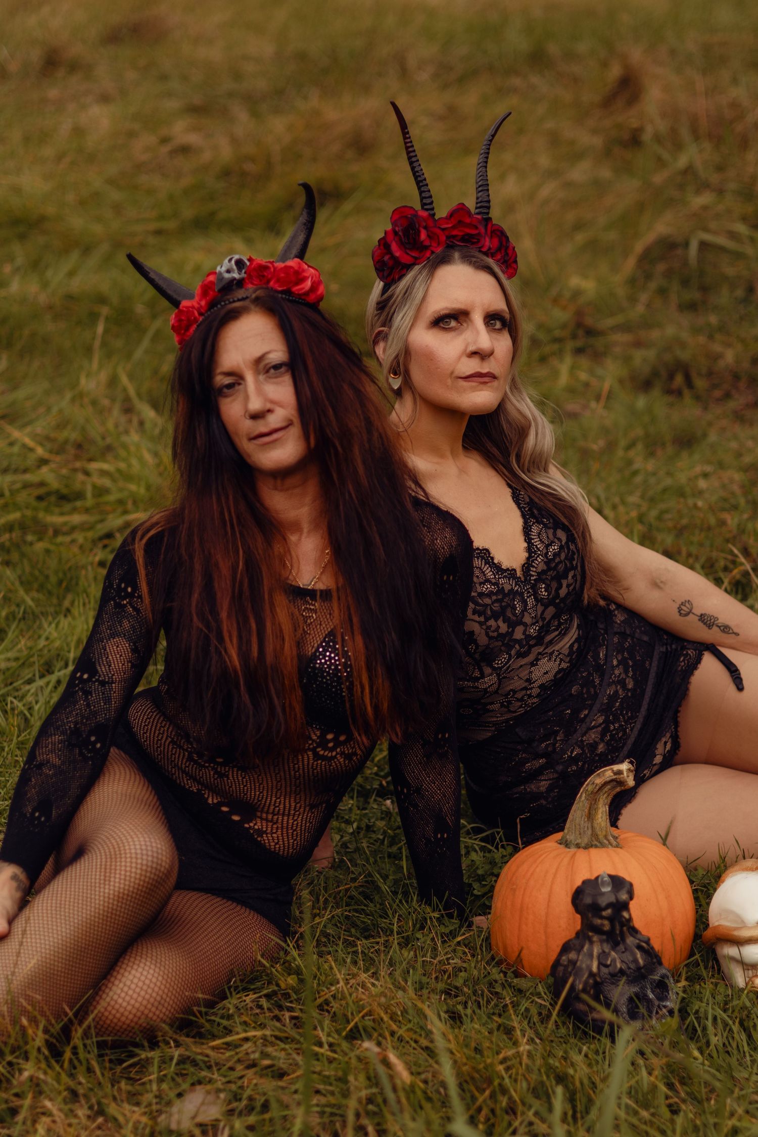Two women in black lace costumes with devil horns and red flower crowns pose outdoors with pumpkins for Halloween.