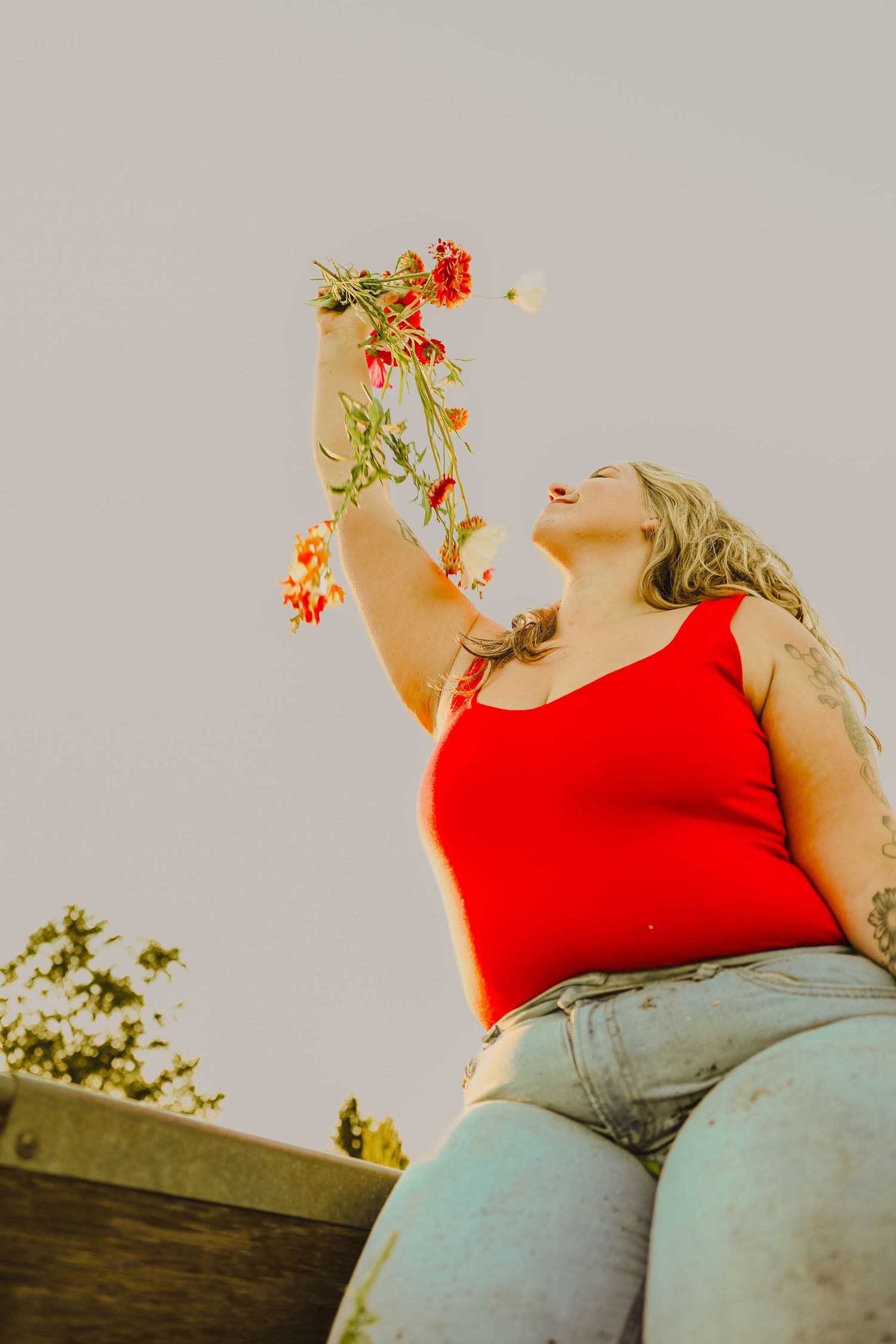 Woman in red tank top and jeans holds red flowers overhead, laughing joyfully against a clear sky.
