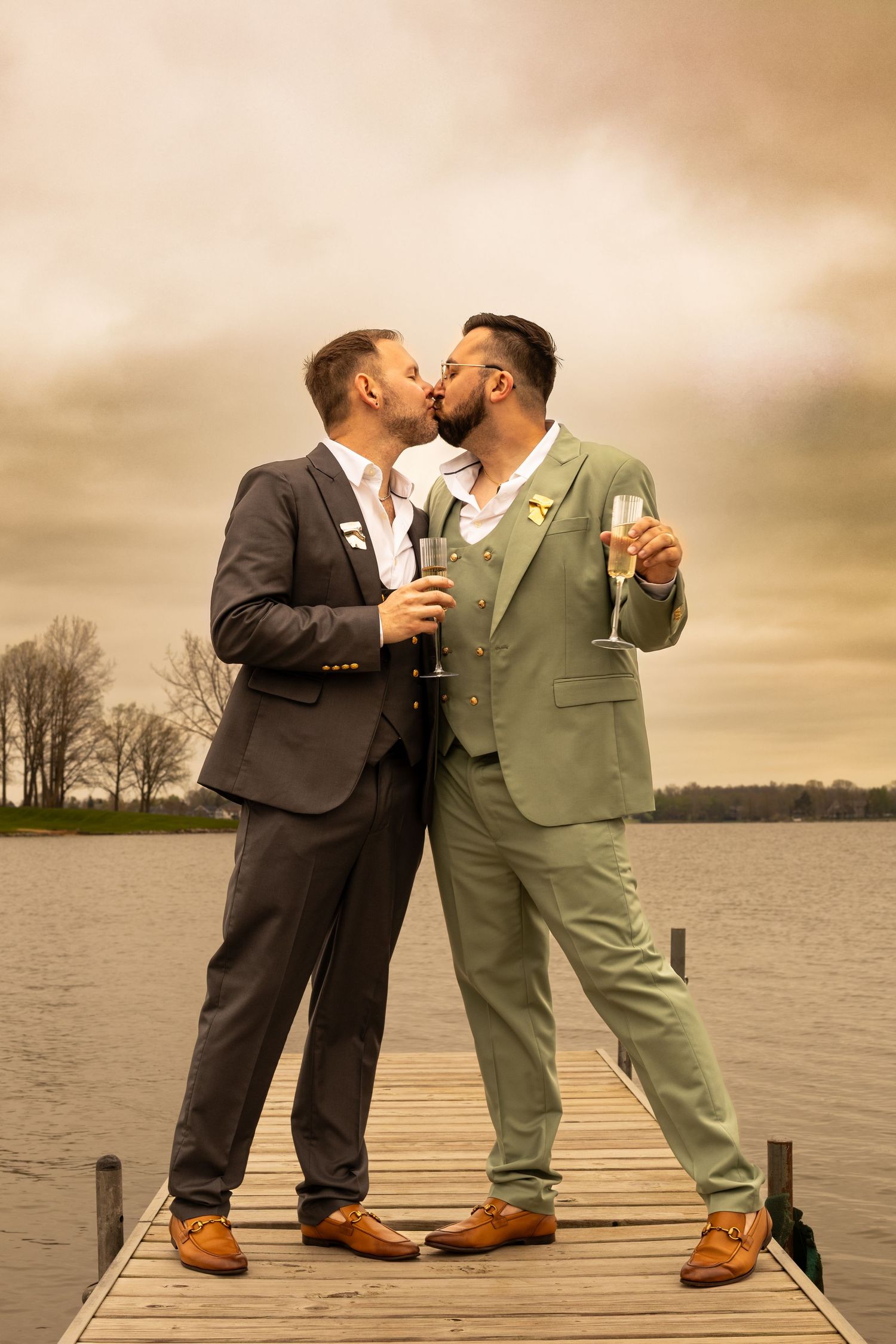 Two grooms in suits kissing on a wooden dock by a lake, holding champagne glasses on their wedding day.