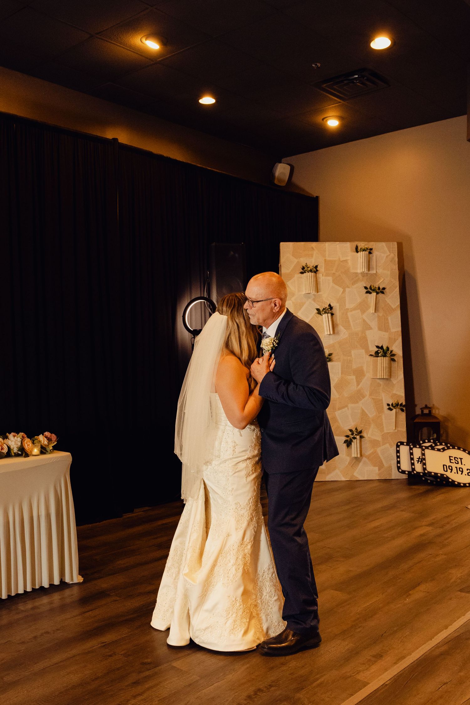 A bride in a veil and ivory gown dances with an older man in a navy suit in a softly lit wedding reception venue.