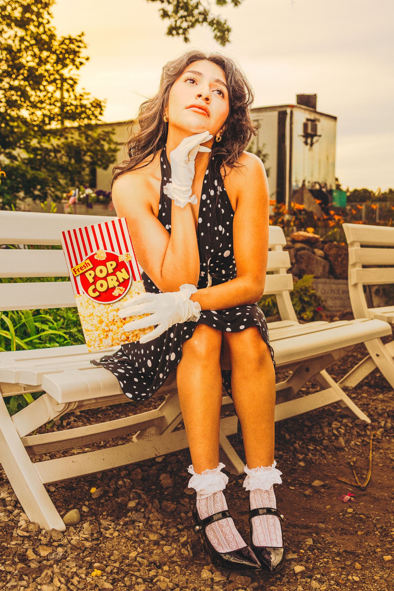 Young woman in polka dot dress holding popcorn bucket sits on white bench outdoors, wearing white gloves and socks.