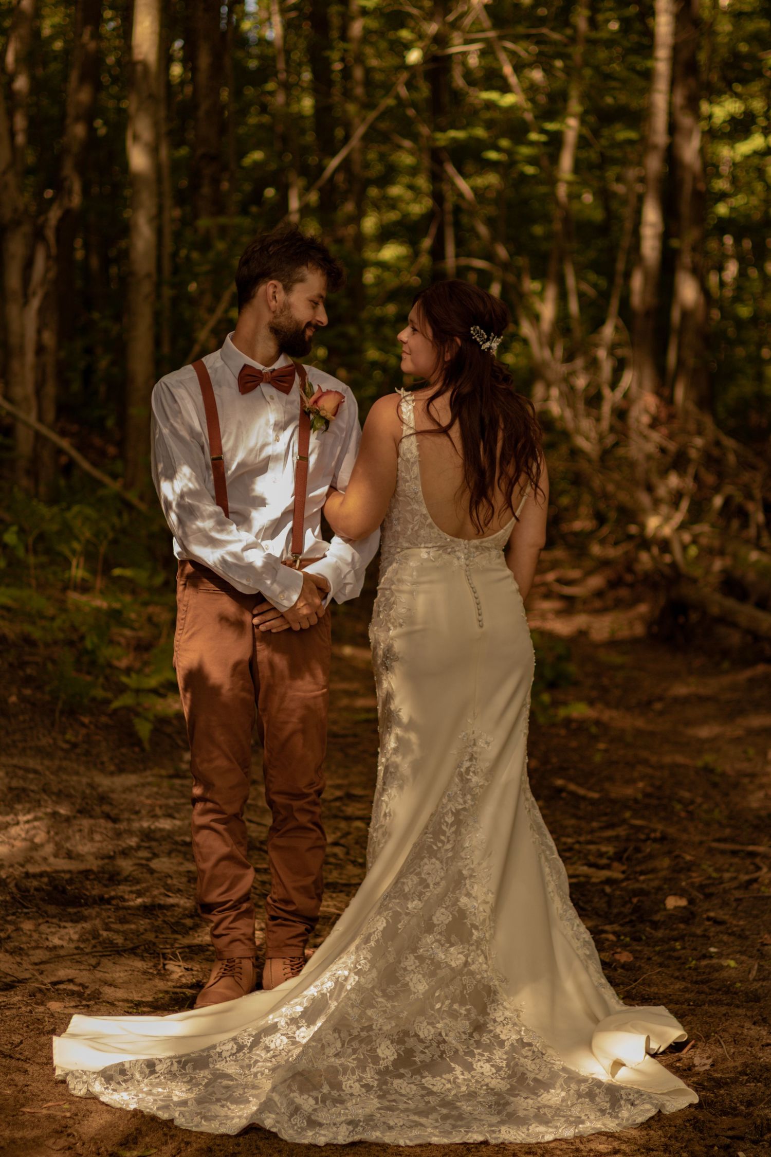 A romantic wedding couple shares intimate moments in a sunlit forest setting, her lace dress trailing beautifully on the ground.