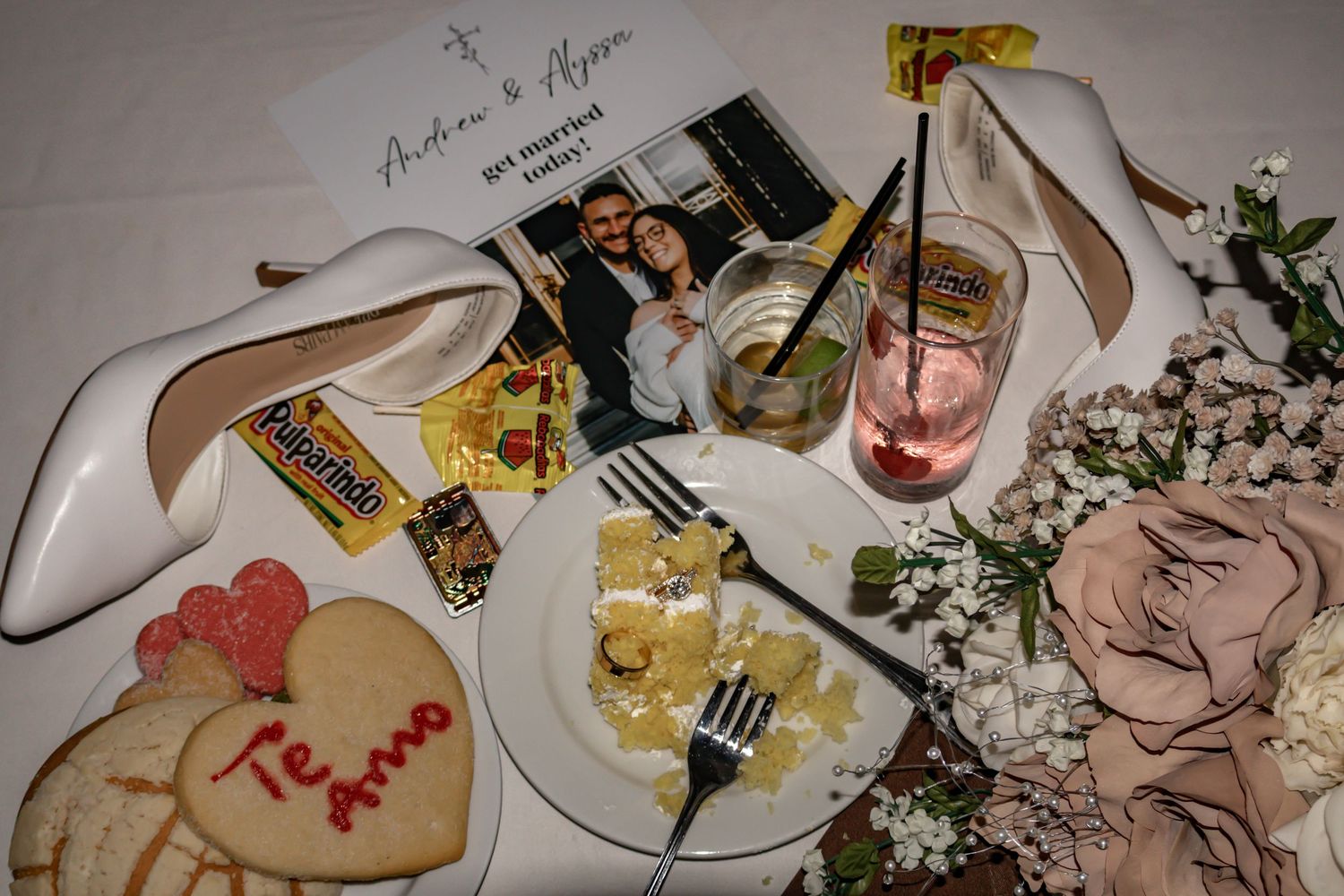 Wedding details including white shoes, heart-shaped cookie, and floral decorations arranged on a light surface.
