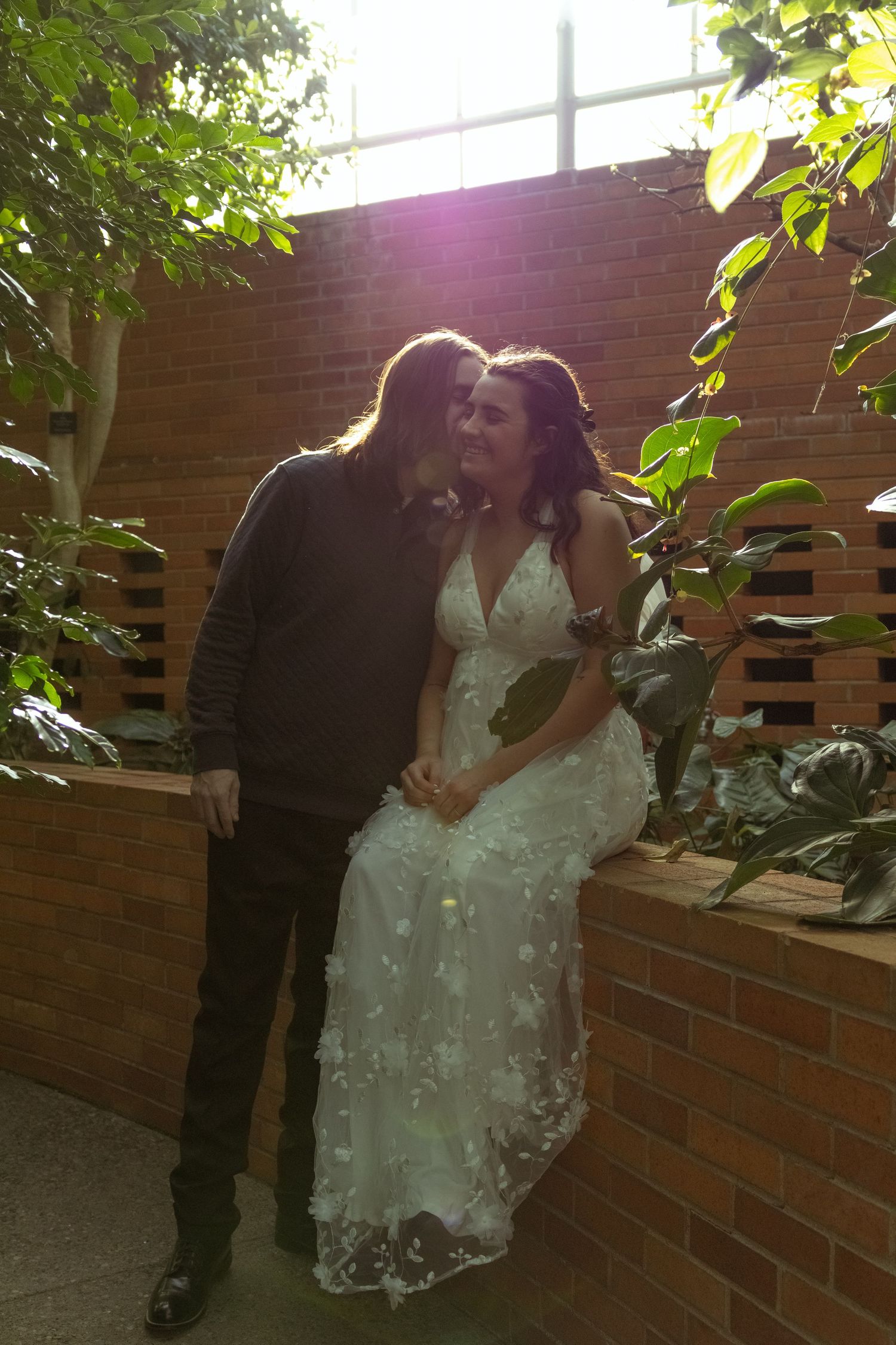 Couple sharing a romantic kiss in a sunlit brick courtyard, bride in white lace gown surrounded by lush green plants.