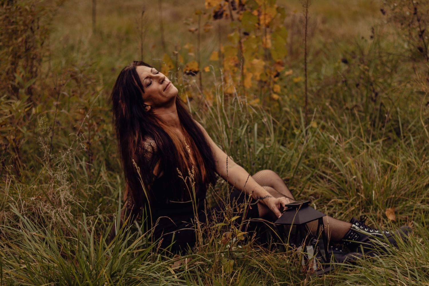 Woman in black dress sitting in tall grass field during autumn, head tilted back with long dark hair flowing.