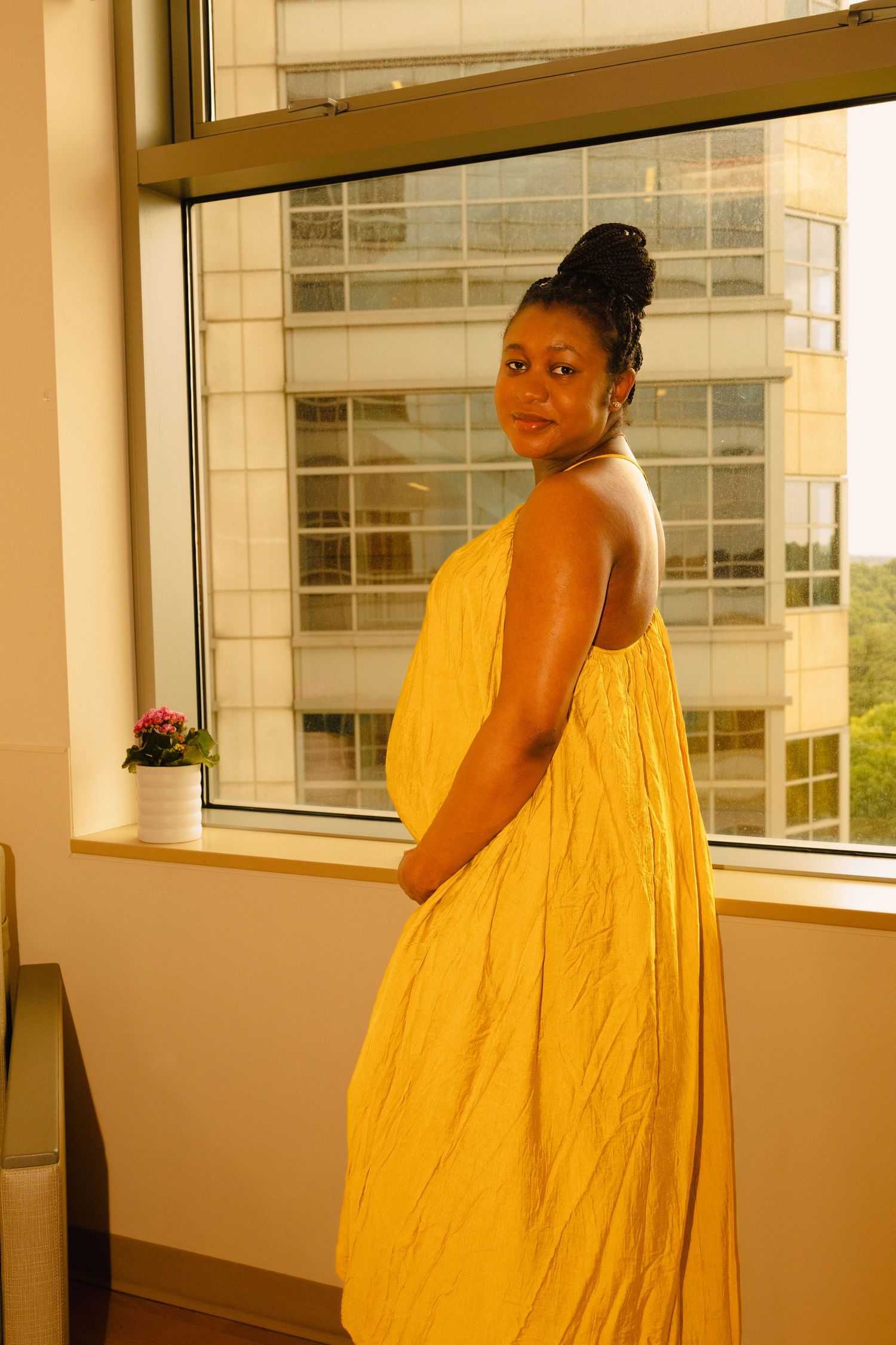 Pregnant Black woman in yellow dress standing by window, looking over shoulder in urban setting.