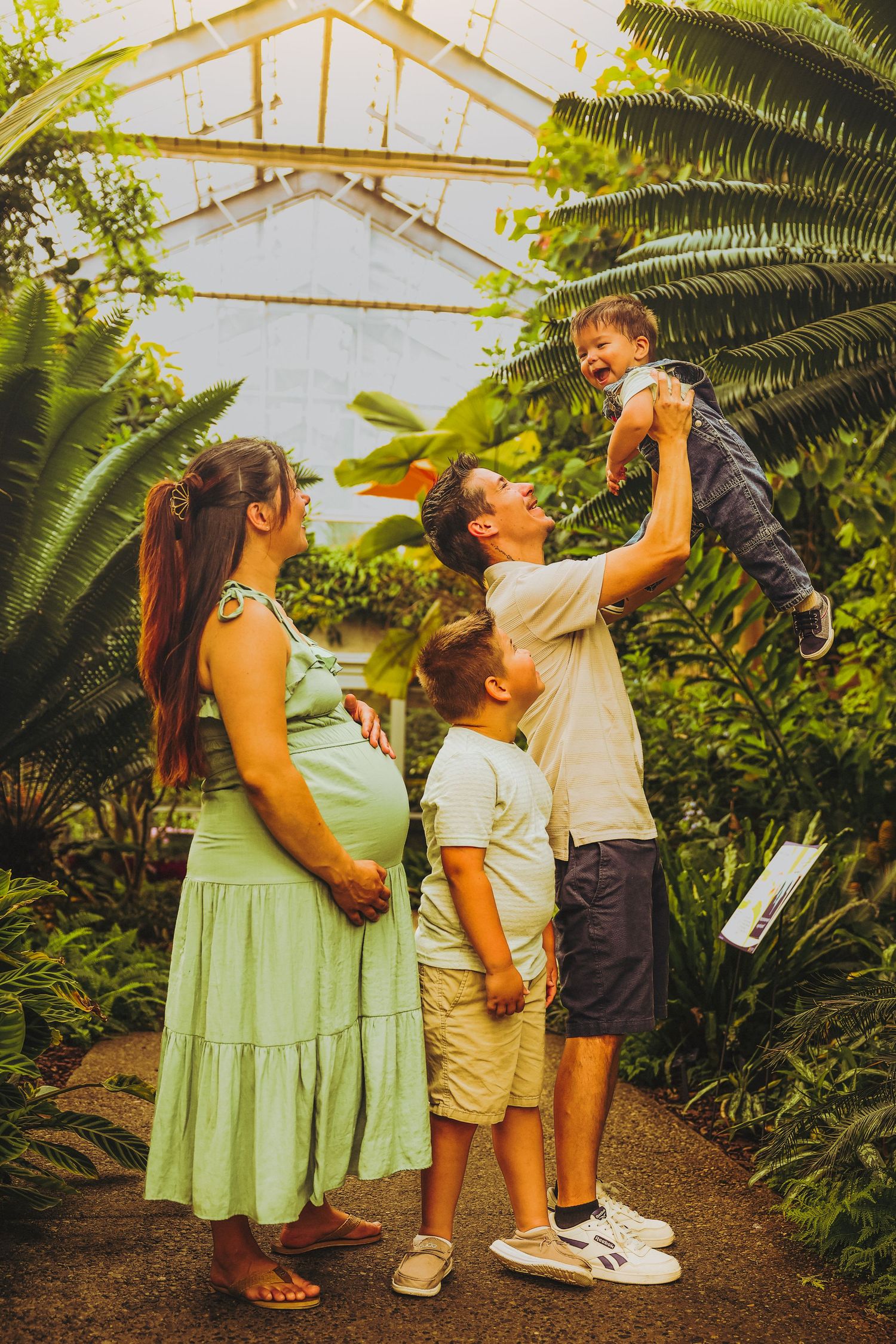 A pregnant woman in a green dress stands with her family in a lush greenhouse as dad lifts a laughing toddler.