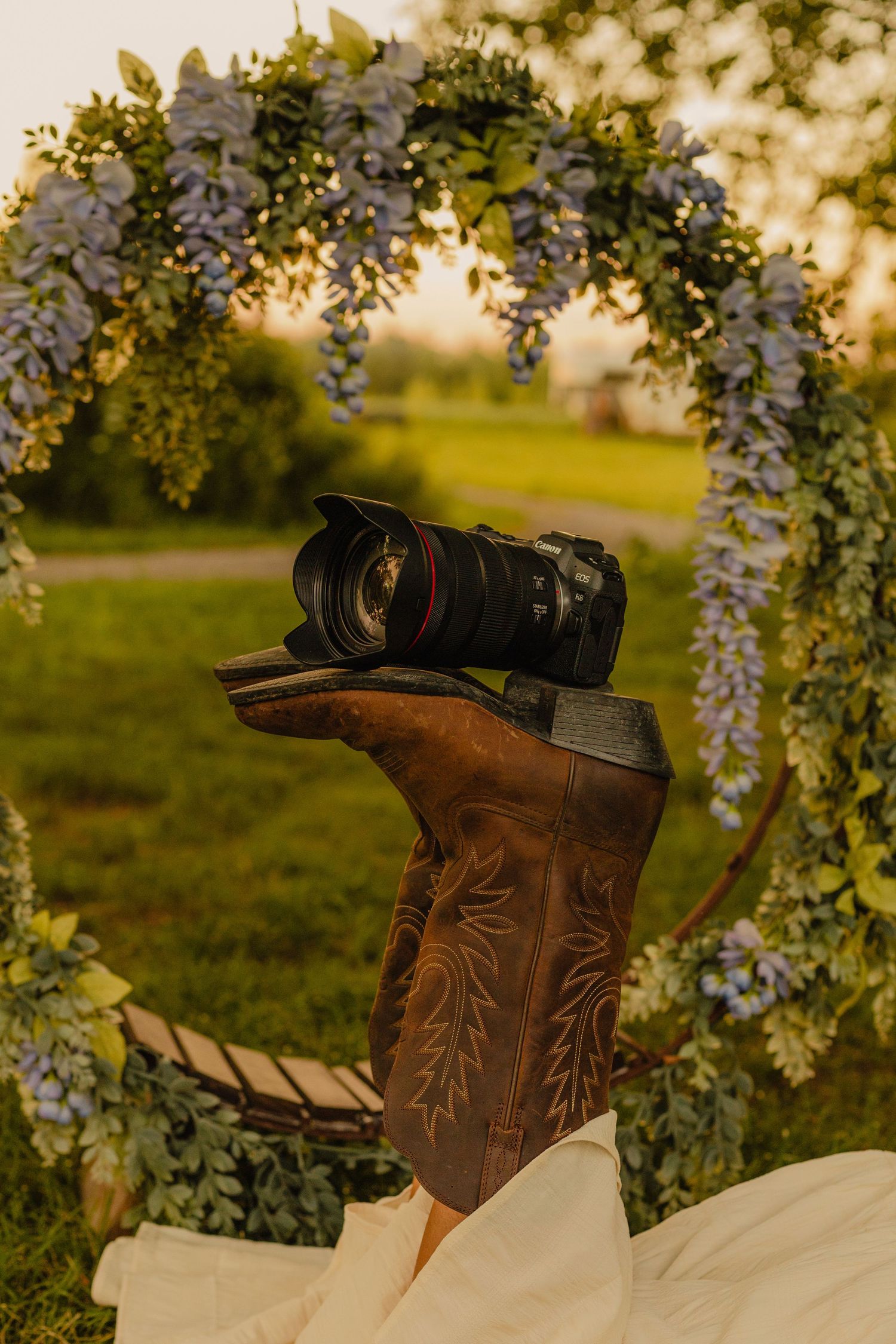 A camera resting on a cowboy boot under a floral arch with wisteria blooms at golden hour.