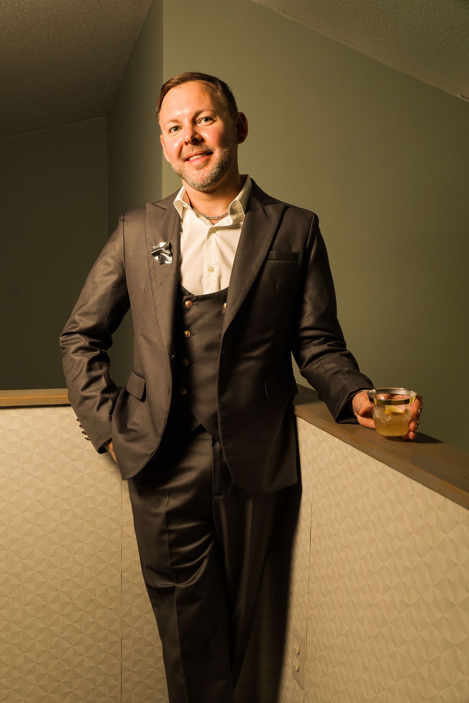 Smiling man in gray three-piece suit holding cocktail glass, leaning against bar counter in elegant venue.