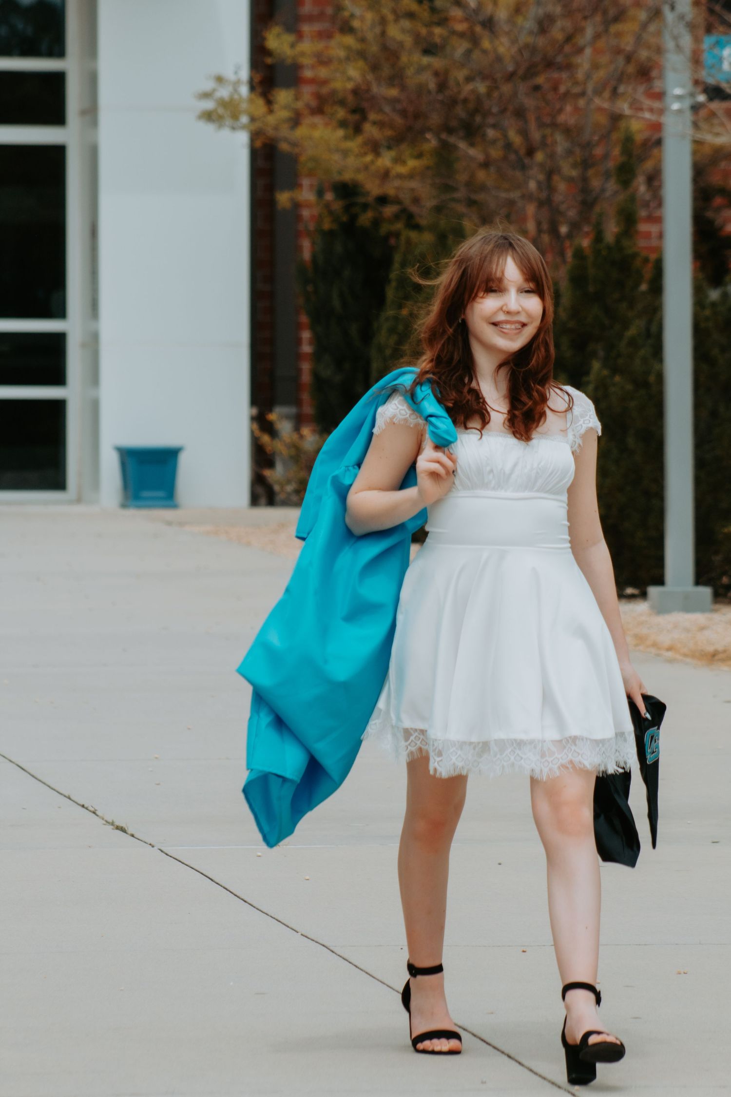 Graduate in white dress carries turquoise gown while walking on campus sidewalk.