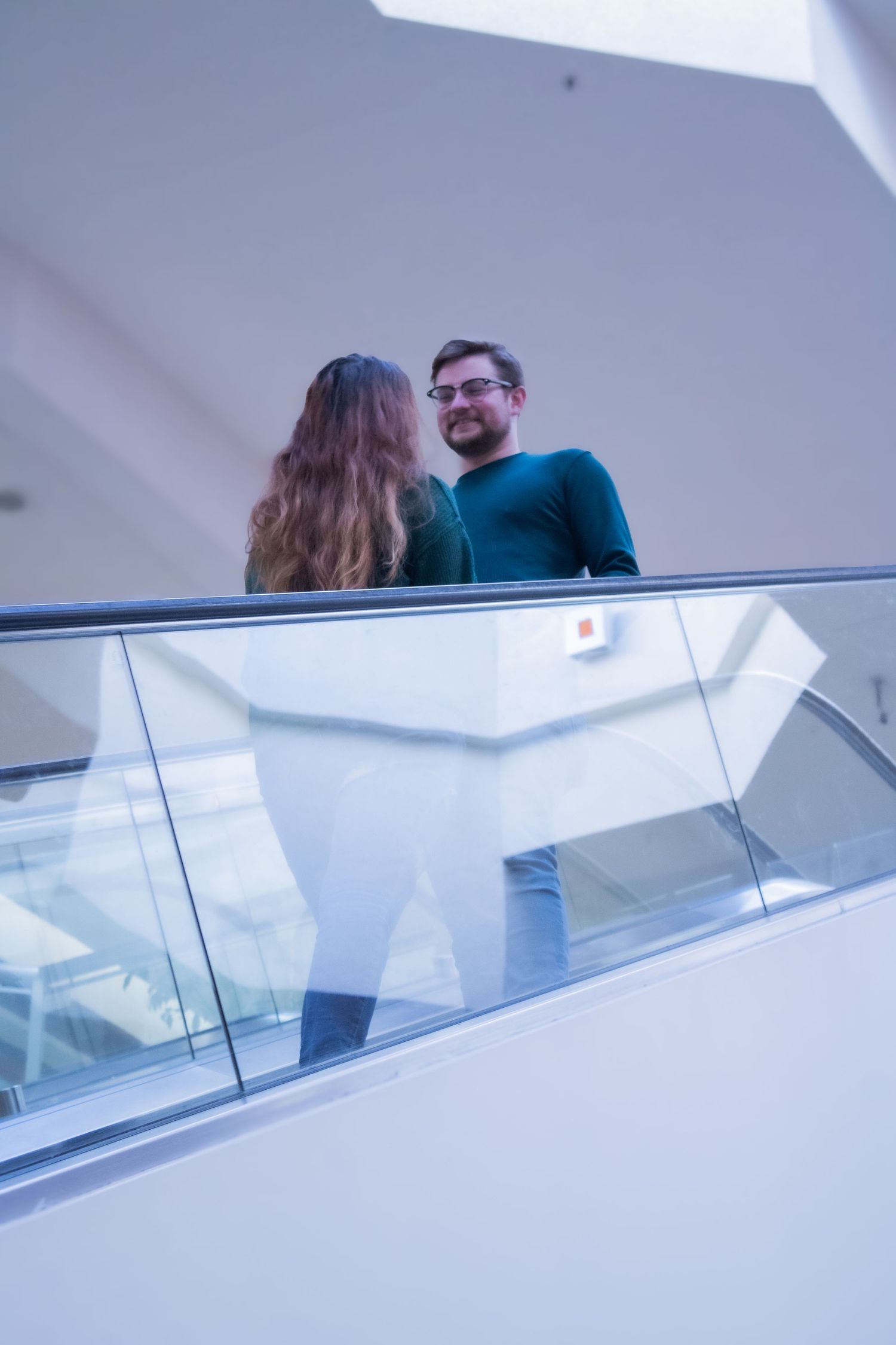 A couple embraces on an elevated walkway with glass railings in a modern interior space.